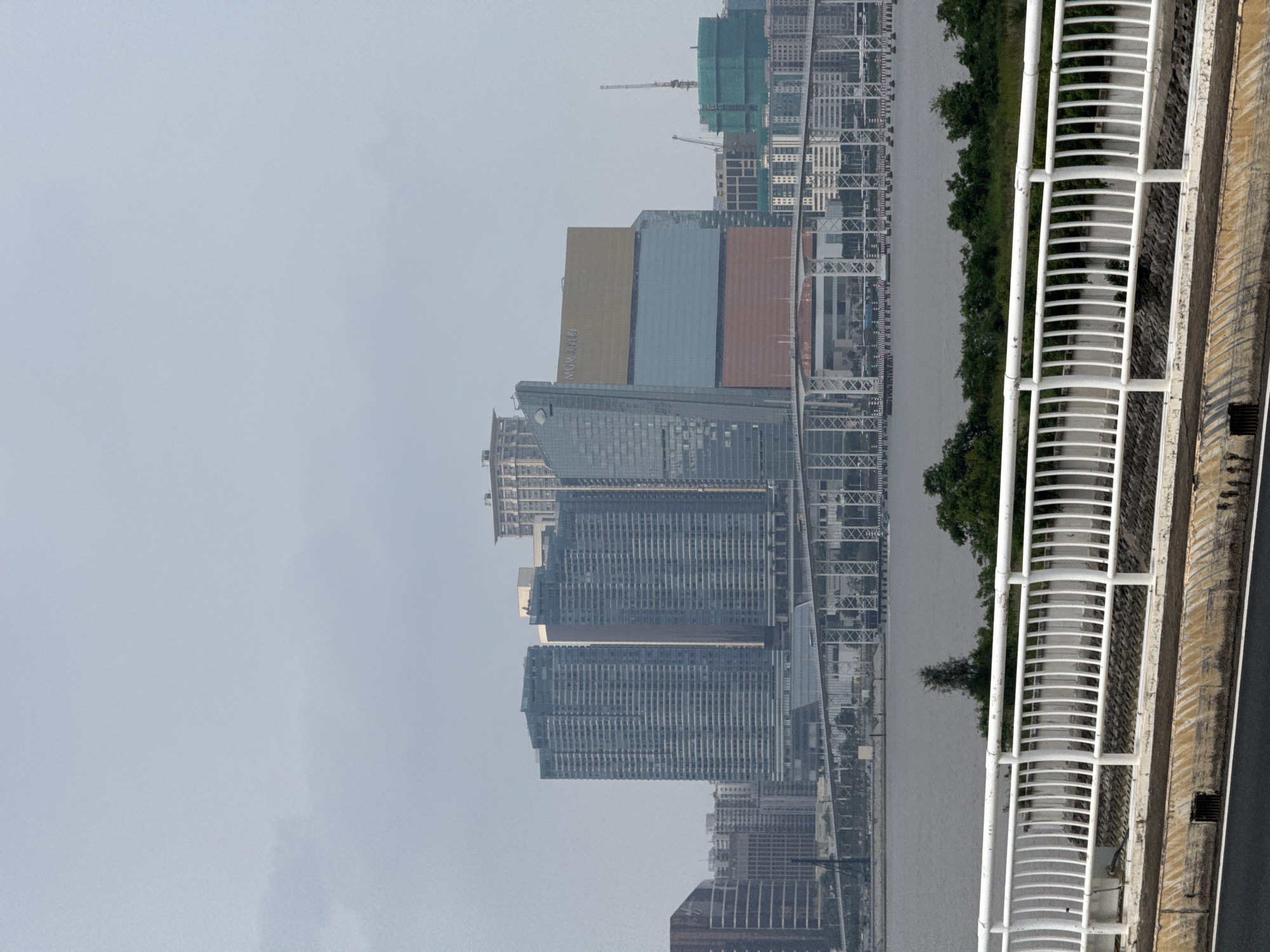 A cityscape, likely in Unknown, Macao, is presented in a rotated view. The city features tall buildings, including skyscrapers and modern architectural structures. The weather appears overcast, casting a hazy tone over the scene. A river runs through the city, reflecting the urban landscape. A bridge or elevated structure with a white railing is visible, suggesting a transportation route. Vegetation, potentially trees or bushes, is situated along the riverbank.