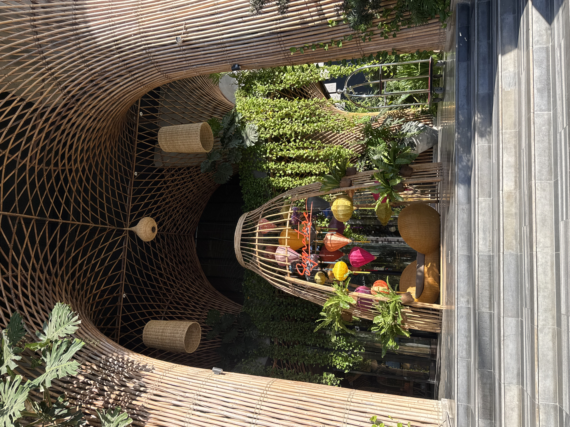 The image depicts the entrance to a restaurant or hotel in Nha Trang, Vietnam, characterized by its extensive use of bamboo architecture and lush greenery. The structure features a curved bamboo roof supported by thick bamboo pillars, creating an inviting, organic archway. Hanging from the ceiling are several woven lampshades and a prominent bamboo cage-like structure in the center, adorned with colorful paper lanterns and a neon sign that appears to spell out "Signature." Cascading vines and various potted plants frame the entrance, adding a tropical feel. In front of the entrance, there are two wicker chairs and a luggage cart. The ground is covered by large, grey tiled steps leading up to the entrance. The scene is brightly lit by natural sunlight.
