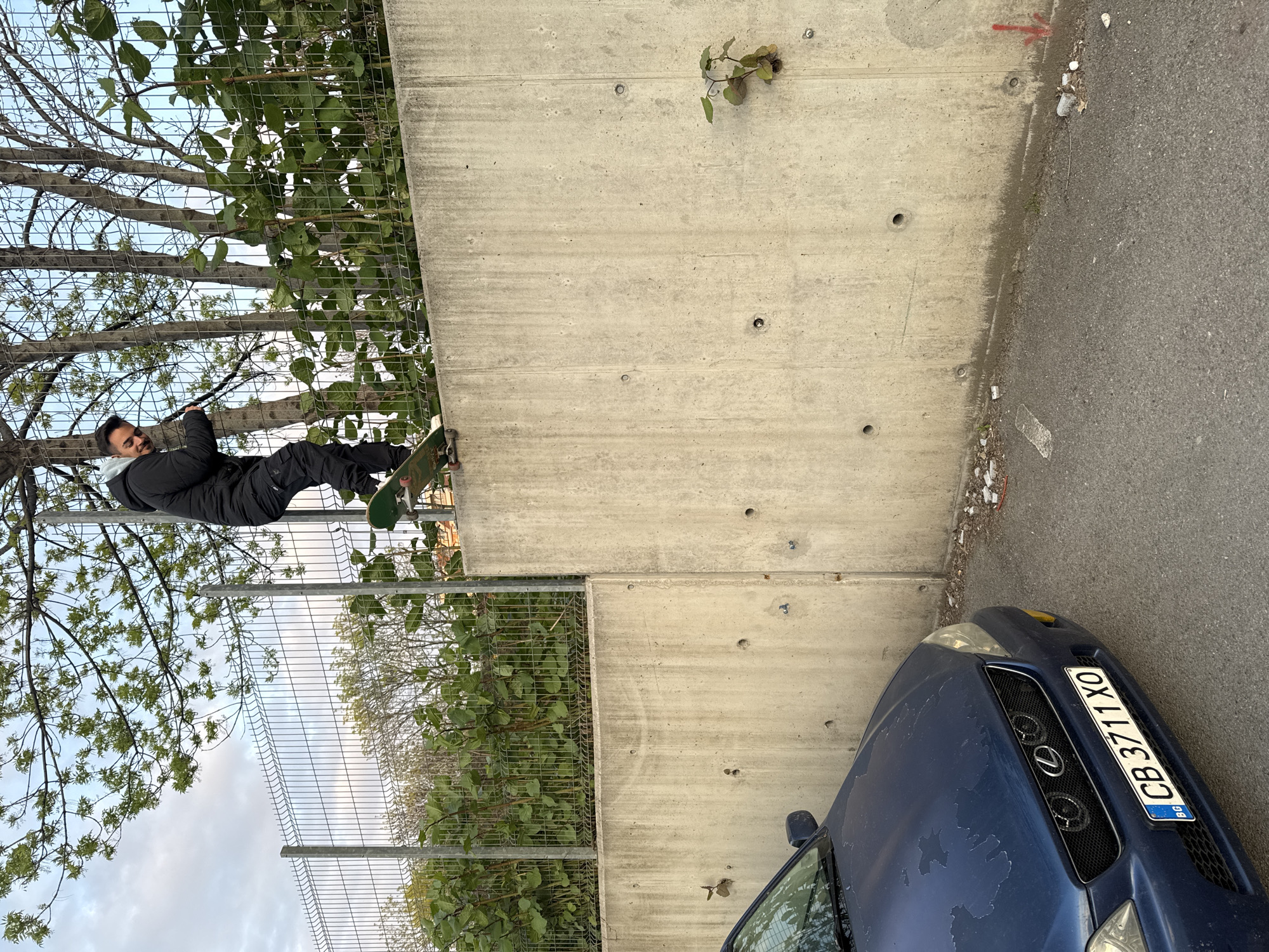 The image depicts a dynamic urban scene featuring a man skateboarding. The man is positioned vertically against a tall, light grey concrete wall, appearing to perform a trick such as a wallride or a balanced pose. He wears a dark jacket over a light hoodie, dark trousers, and has a short beard, displaying a slight smile or look of concentration. A green skateboard is visible under his feet.
To the left of the concrete wall, a metal mesh fence runs alongside a row of trees with green leaves, under a partly cloudy sky. The presence of the fence and trees suggests a boundary or green space adjacent to the concrete structure.
In the lower right foreground, a dark blue car, likely a sedan, is partially visible, parked on an asphalt road next to the concrete wall. The car's hood shows signs of wear, with peeling paint. Its front grille features an "L" emblem (likely Lexus), and the license plate clearly displays "BG CB 3711 XO", with "CB" indicating Sofia, Bulgaria, consistent with the provided location context. The asphalt surface next to the wall is littered with some debris, and a faint red arrow is visible on it.
The lighting suggests it is daytime, with soft, diffused light indicating either an overcast sky or late afternoon conditions. The overall setting is an urban environment, capturing a moment of an individual engaged in skateboarding.