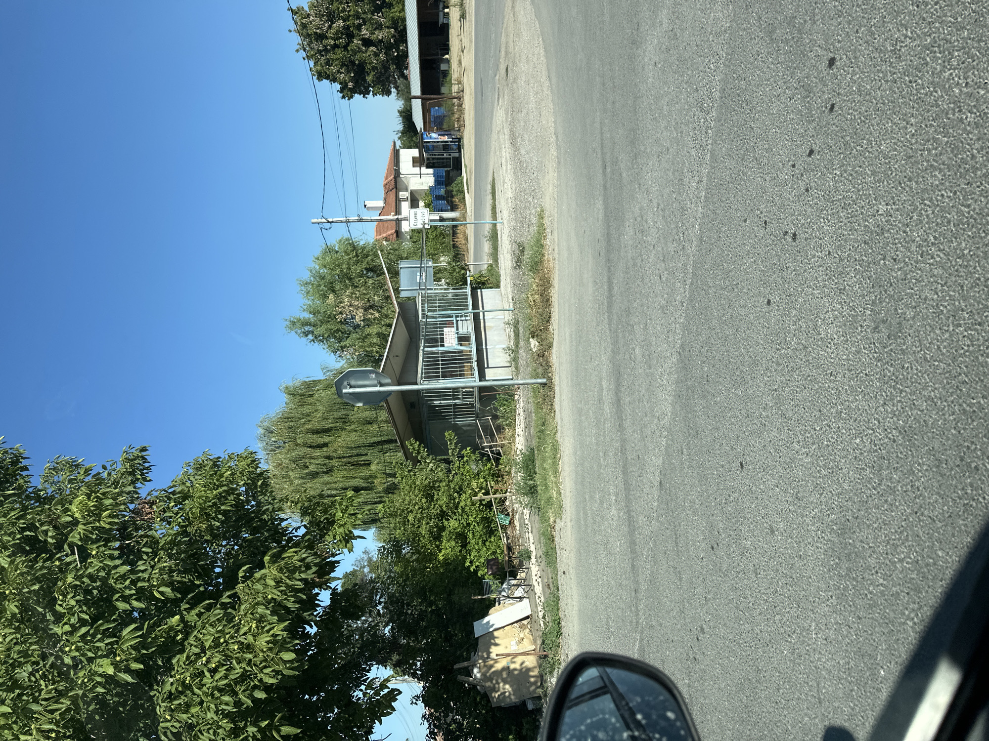 The image captures a street scene in the city of Kadievo, Bulgaria, under a clear blue sky. In the foreground, a paved road occupies the lower portion of the frame. To the left, a car's side mirror is visible, reflecting the surrounding environment.
Dominating the mid-ground is a small, open-sided structure, possibly a kiosk or bus stop shelter, with a metal railing around its perimeter. Behind it, dense green foliage, including trees and bushes, creates a natural backdrop. A tall, slender tree with drooping branches stands to the left of the kiosk, while a larger, leafy tree is positioned further back. Various discarded items, including wooden planks and what appears to be debris, are piled near the left side of the structure.
Further in the background, a residential building with a tiled roof is visible. To the right of the kiosk, a street sign with text, likely indicating the location, is planted in the ground. Beyond that, a long, low-slung building with a corrugated roof stands, possibly an outbuilding or workshop. Electrical wires stretch across the sky, supported by a utility pole. The overall environment suggests a rural or semi-rural setting within the town.