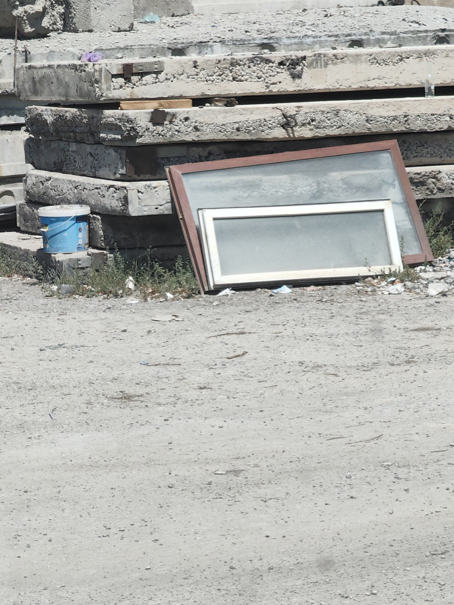 The image shows a pile of concrete slabs and a window frame leaning against them in an outdoor urban setting. A blue bucket sits on a smaller concrete block at the base of the pile. The ground is covered in dirt and small stones, with some scattered debris and vegetation. The scene appears to be a construction or demolition site, possibly in the city of Shymkent, Kazakhstan, given the context provided. There are no people or animals visible in the image, and no discernible actions are taking place. The overall impression is one of neglect and disuse.