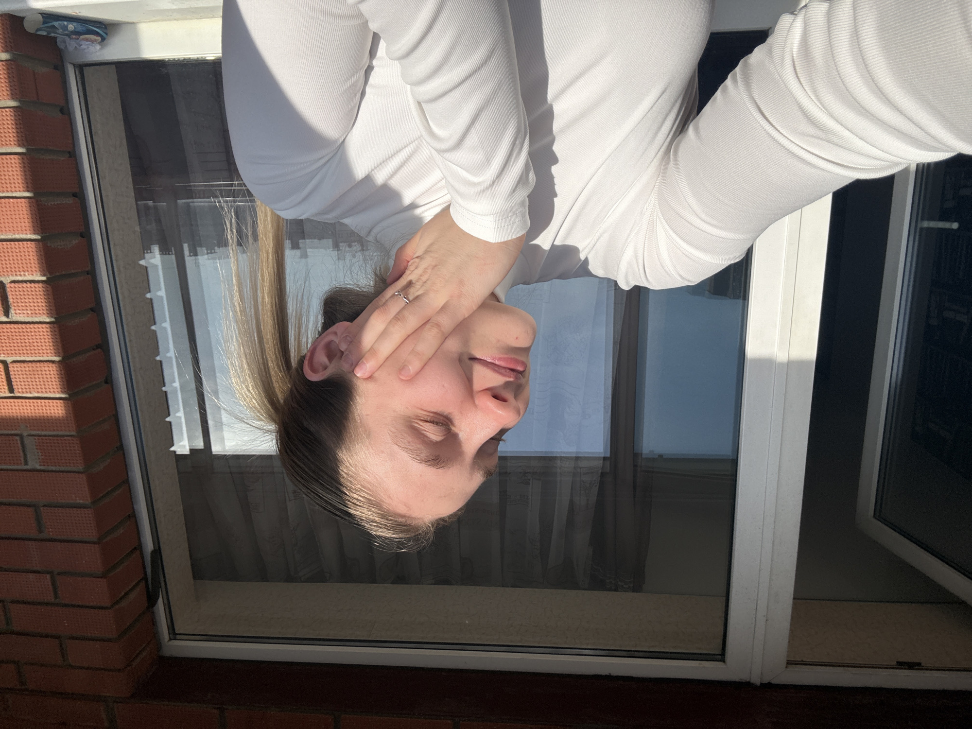 A young woman is seen in an upside-down perspective, her face partially obscured by her hand. She's wearing a white long-sleeved top and a ring is visible on her finger. She is positioned against a window frame, with a brick building visible outside. The reflected sky is visible in the window panes. Based on available data, it is not possible to confirm the location of this picture is the city of Izhevsk, Russia.