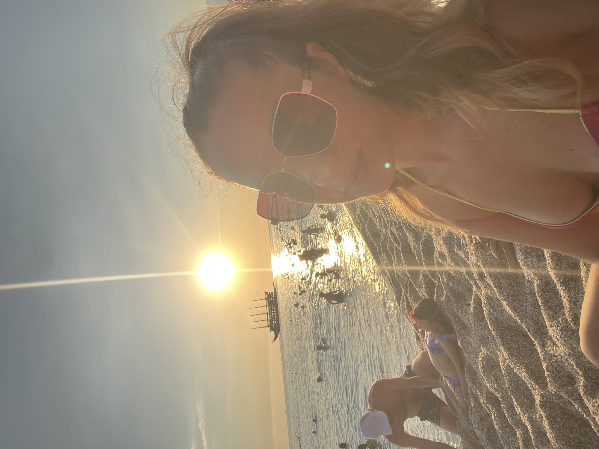 The image captures a sunny day at a beach in Alanya, Turkey. The sun is low in the sky, suggesting it is either early morning or late afternoon, casting a warm, golden light across the scene. The water is calm and reflects the sunlight, creating a shimmering effect. In the distance, a traditional sailing ship with multiple masts is visible on the water.
In the foreground, several people are present. A woman wearing sunglasses is taking a selfie, with her face partially visible. Her hair is light brown and falls around her shoulders. She appears to be wearing a pink bikini top. To her right, a child wearing a purple and blue swimsuit and pink goggles is sitting on the rocky shore, looking towards the sea. Further to the right and slightly out of focus, a person wearing a white hat and patterned swim trunks is also visible on the shore. The beach appears to be made of pebbles.
The overall mood of the image is relaxed and summery, with the warm sunlight and the presence of people enjoying the beach environment. The vibrant colors of the swimwear and the natural beauty of the beach and sea contribute to this atmosphere. There is no visible text in the image.