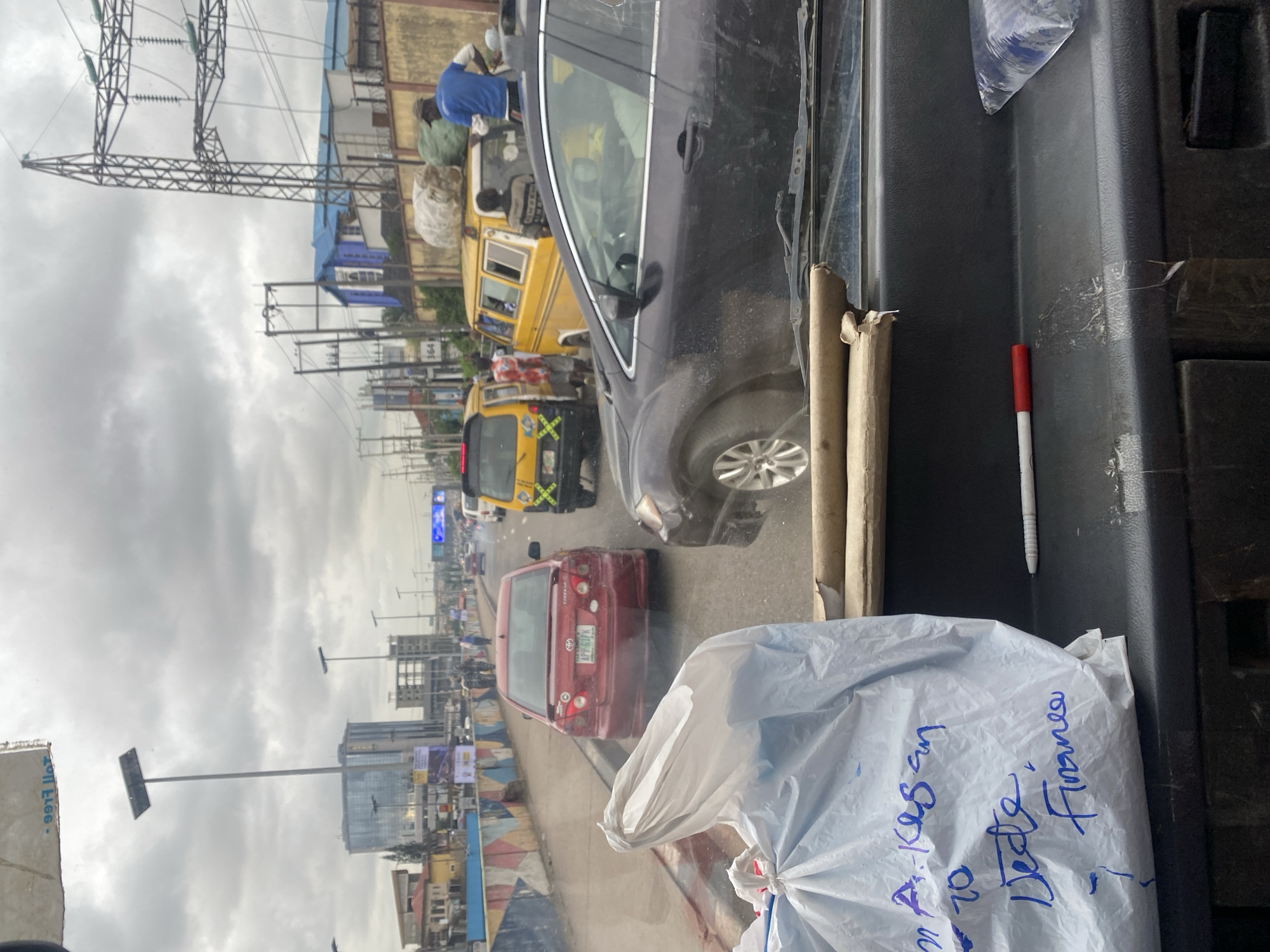 The image is a street-level view of traffic congestion in Ikeja, Nigeria, likely taken from inside a vehicle. The sky is overcast. In the foreground, a white plastic bag with blue handwritten text is visible on the dashboard, along with a red-tipped pen. The bag has writing that appears to say "A. Kesan to Dede, Finance".
The street is lined with various vehicles, including a red Toyota sedan with license plate "APP 61FK", a dark-colored sedan, a yellow minibus, and another yellow minibus behind it. There are also power lines and utility poles along the side of the road. In the background, buildings of varying heights and architectural styles are visible, including a modern glass-fronted building and older structures. Several people are present, some standing or walking near the vehicles. One person in a blue shirt is standing on top of a yellow minibus, seemingly overseeing goods or passengers. The overall scene depicts a busy urban environment experiencing traffic delays.