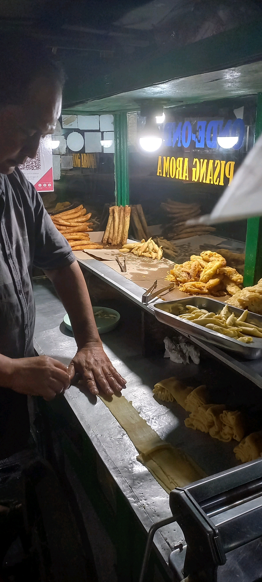 A man, likely a vendor, is seen in the foreground of this image preparing food at a street-side stall in Teluk Dalam, Indonesia. He is wearing a dark grey collared shirt and is using both hands to flatten a thin piece of dough on a metal counter. Behind him, a display of fried foods is visible, including what appear to be fried bananas and other assorted fried items, some piled in metal trays and others on paper. A sign with text is visible in the background, with yellow lettering that reads "AMOMA PISANG" and blue lettering above it. The stall appears to be an outdoor establishment, illuminated by artificial lighting, suggesting it is nighttime or a dimly lit indoor space. The overall environment suggests a food preparation and sales setting typical of a night market or street food vendor in the city.