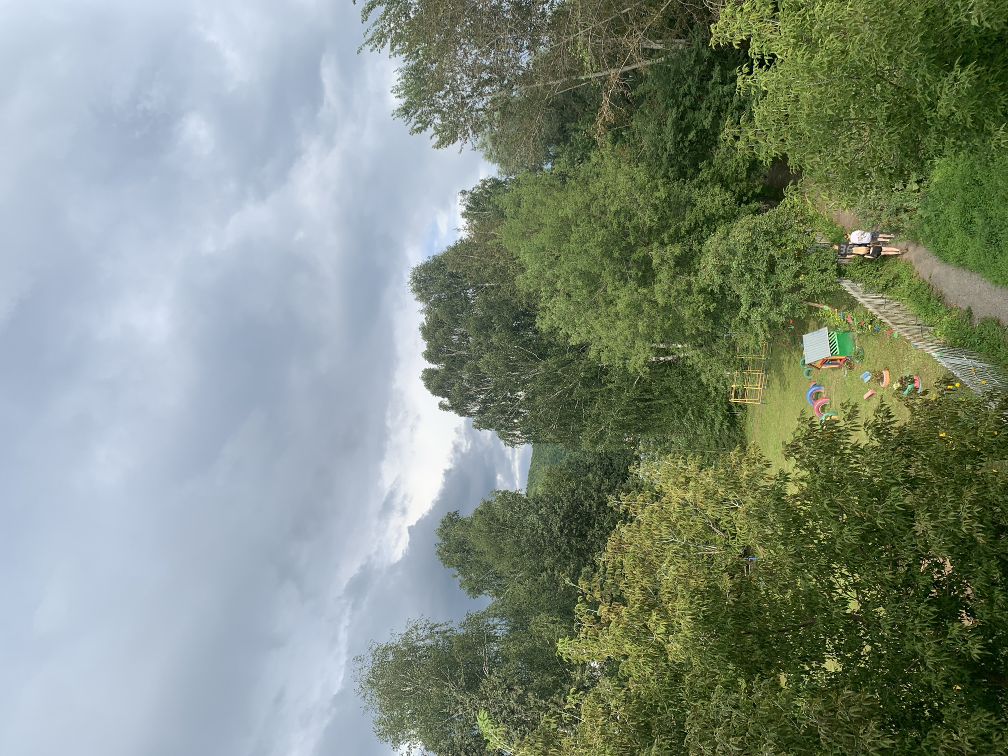 This image captures a view from above of a park-like area on a cloudy day. In the foreground, lush green trees with dense foliage dominate the scene. To the right, a dirt path winds through the grass, bordered by a white picket fence. Two individuals, appearing to be adults, are walking away from the camera on this path.
Further into the park, there is a small, brightly colored playground area. Elements include a small, multi-colored structure resembling a playhouse, colorful rings laid out on the grass, and a yellow metal play structure with swings. The background is filled with a dense forest of trees, with hills visible in the distance. The sky is overcast with heavy, dark clouds, suggesting an impending storm or recent rainfall. The location is identified as Asha, Russia, implying this is a public park or recreational space within the city.