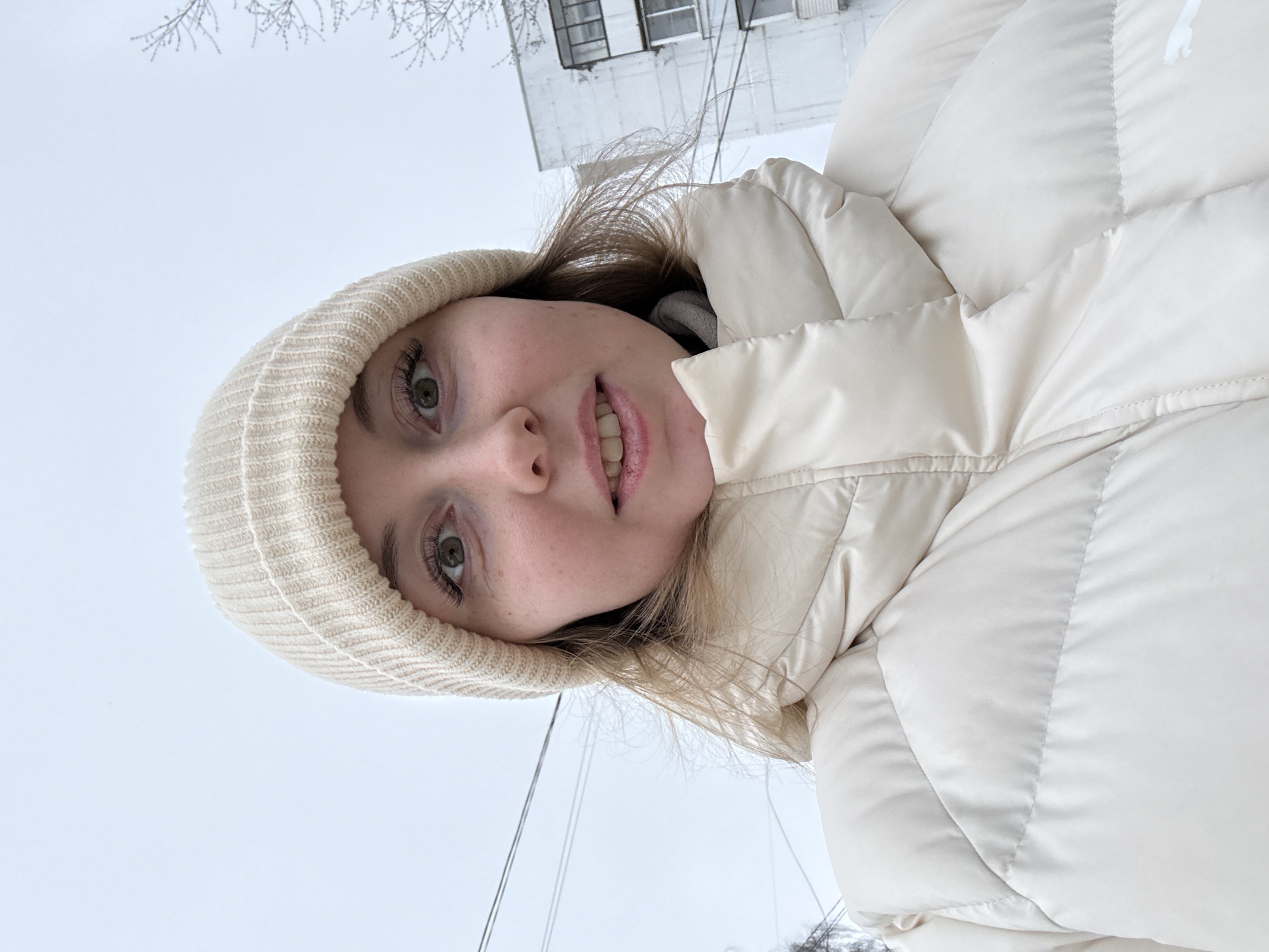 A young woman is wearing a white beanie and a puffy white jacket while standing outdoors in front of a building. The sky is overcast and gray. Bare tree branches are visible in the background.
