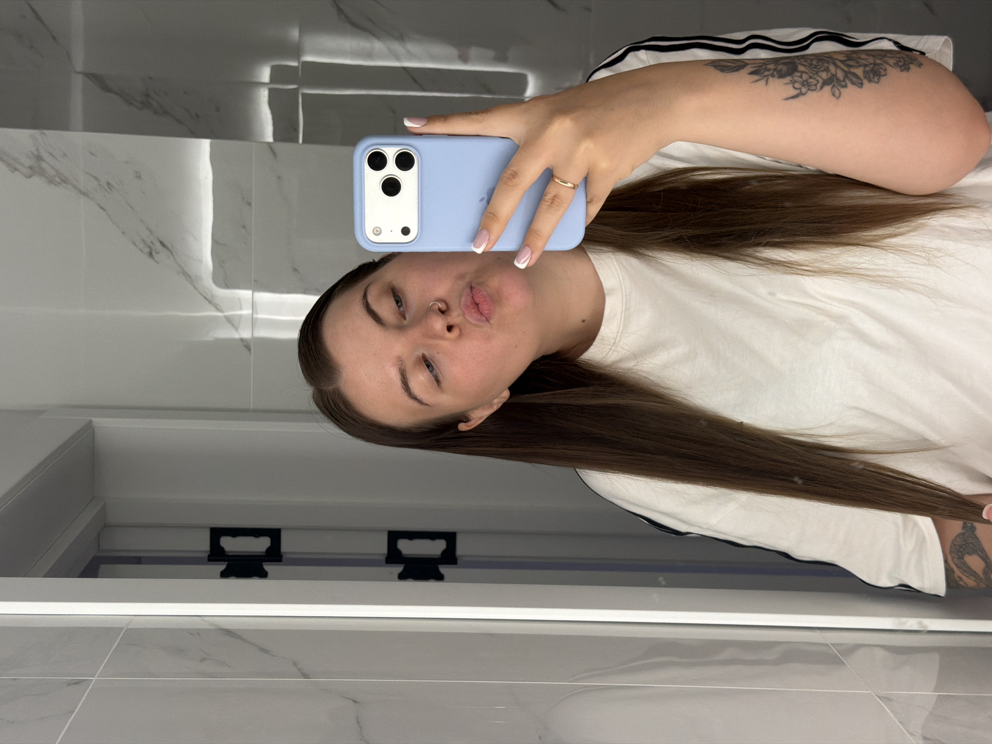 A woman is taking a selfie in a bathroom with tiled walls. She is holding a light blue iPhone with a triple-lens camera. The woman has light skin, brown hair, and a nose ring. She is wearing a white t-shirt with black stripes on the sleeves. Her right arm has a floral tattoo. She is making a kissing face. The location is likely Chaikovsky, Russia, based on image analysis.