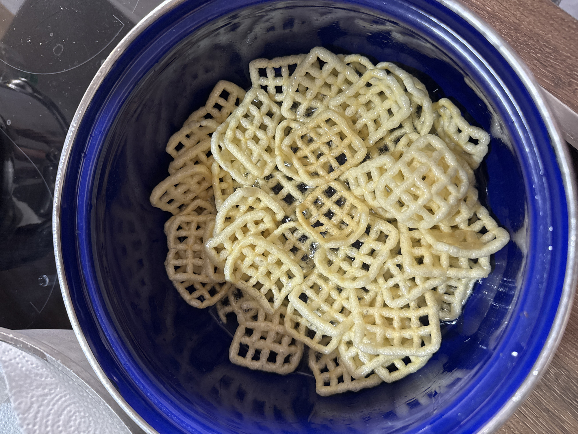 A blue ceramic bowl filled with golden waffle-shaped potato chips is placed on a wooden surface. The chips are stacked unevenly, with some overlapping. A small amount of oil or liquid is visible at the bottom of the bowl, surrounding some of the chips. In the background, out of focus, is a glimpse of a stovetop and a white paper towel in a metallic container. The lighting is bright, casting subtle shadows.