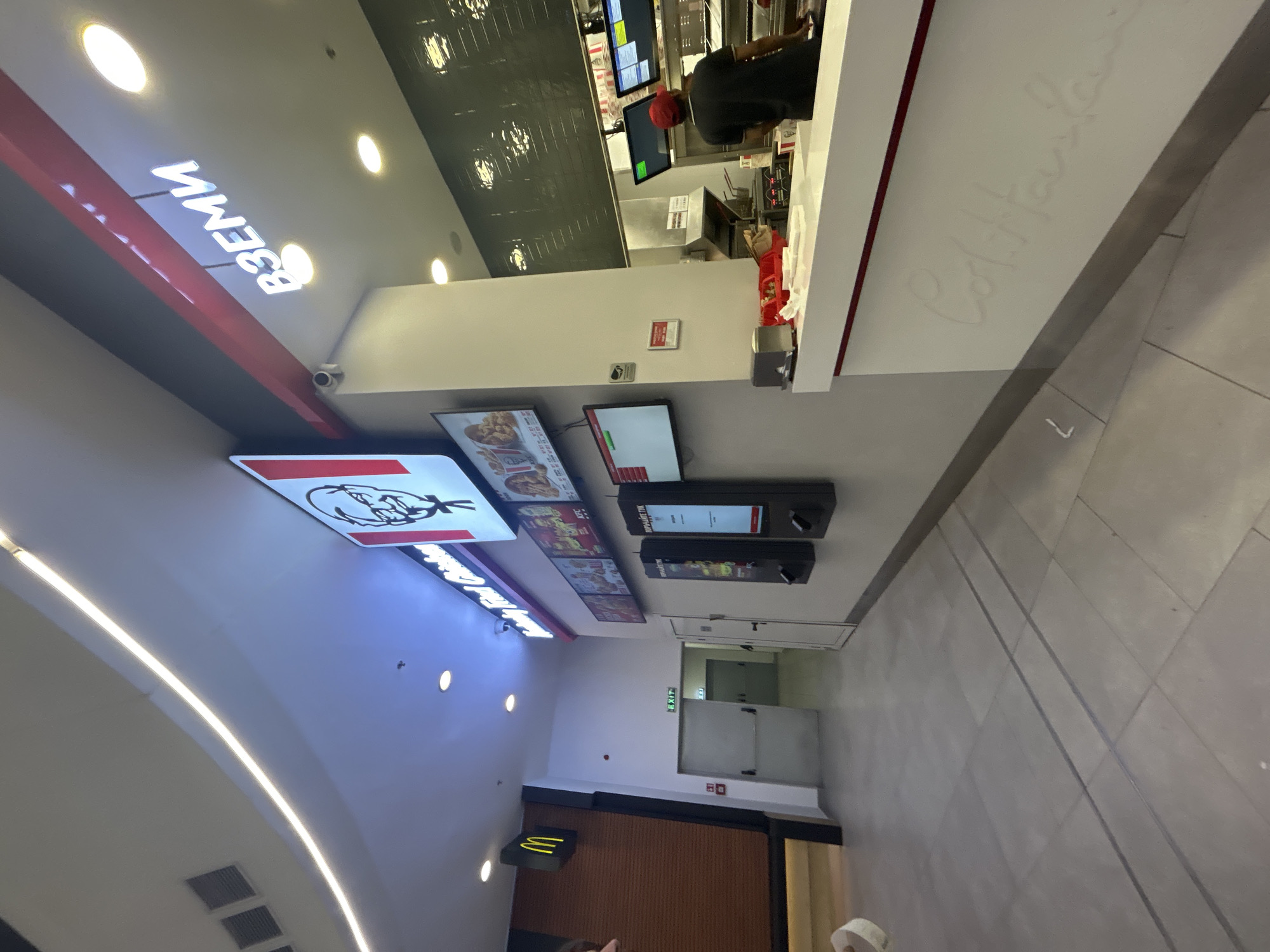The image shows the interior of a KFC restaurant. In the foreground, a white counter with a red stripe is visible. The words "Colt. Haslam" are embossed on the counter in cursive. Behind the counter, an employee wearing a red cap and a dark shirt is preparing food. Various screens and kitchen equipment are visible in the background. To the left of the counter, a hallway leads to an exit door with an "EXIT" sign. On the wall above the counter, a KFC logo and menu boards are displayed. Further to the left, a McDonald's logo is mounted on a wall. The overall environment appears to be a modern indoor commercial space, likely a food court or a standalone restaurant. The lighting is a mix of overhead fluorescent lights and accent lighting. The floor is tiled.
