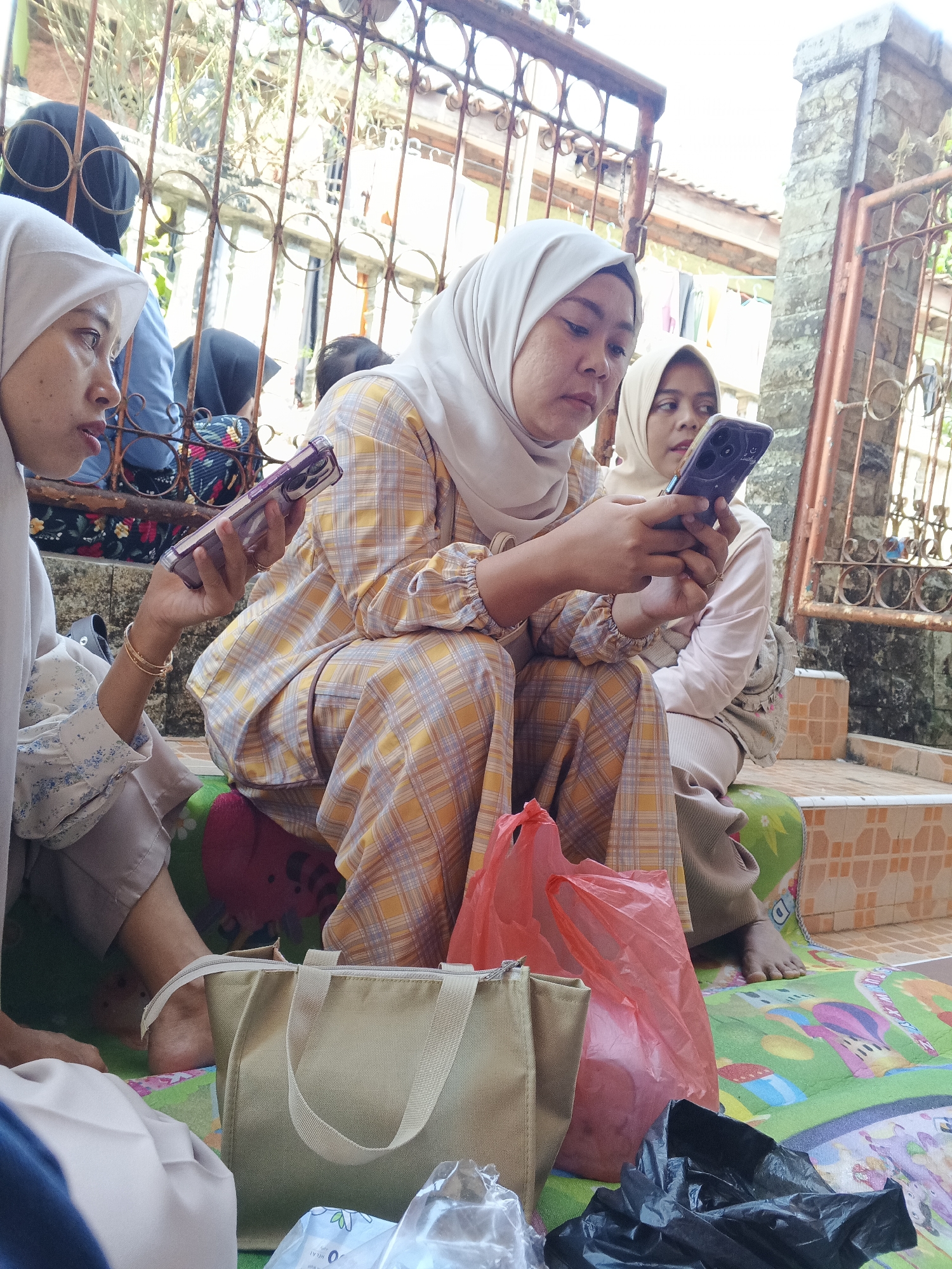 In Warungkiara, Indonesia, three women are seated on a mat outdoors. The woman on the left wears a light grey hijab and a patterned blouse. She holds a smartphone in her left hand, with a bracelet on her wrist. The woman in the center wears a yellow and white checkered hijab and a matching outfit. She is looking at her dark purple smartphone, which is held in her hands. To her right, another woman wears a light beige hijab and is partially visible, also looking at a smartphone. In front of them are two plastic bags, one pink and one red, and a beige tote bag. The background shows a metal fence and a stone wall, suggesting an outdoor setting adjacent to a building.