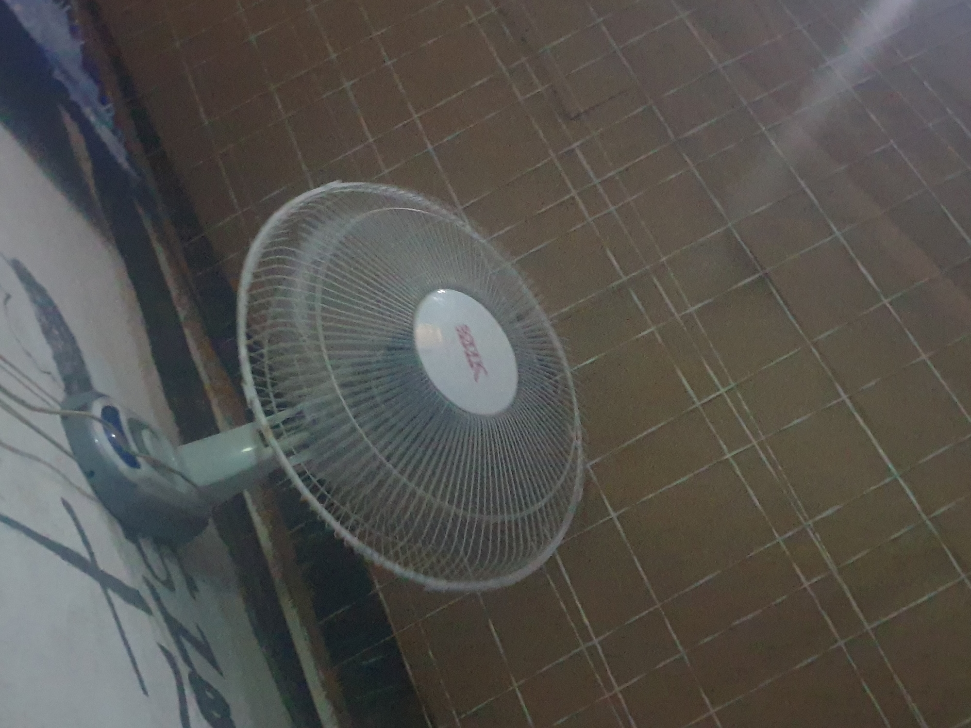 A wall-mounted fan is angled towards the viewer, its white grille covering the central portion of the image. The fan has a white circular center with a red logo or text. The fan is attached to a light gray bracket and then to a wall, which is white and appears to have some dark markings or graffiti on it. The fan is positioned in front of a brown tiled floor. The lighting in the scene is dim, casting shadows and creating a slightly blurry effect, particularly on the fan's blades. There are no people visible in the image. The scene appears to be indoors. There is no information in the image to suggest it is located in Washington, United States of America.