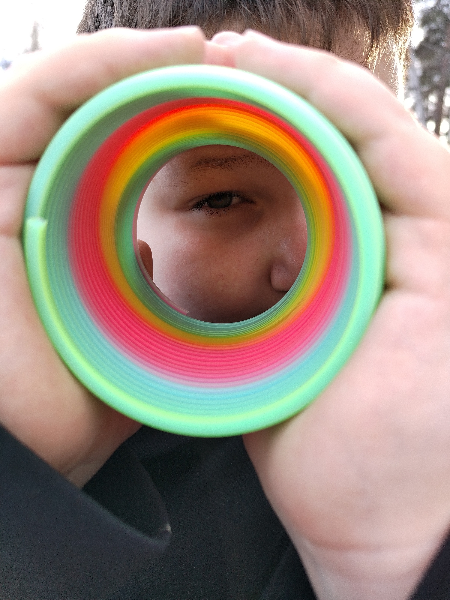 A person's face is seen through the center of a coiled, rainbow-colored toy spring. The spring, held by two hands, is predominantly green with bands of red, orange, and yellow. The person's left eye and part of their nose are visible through the spring's opening. The person has brown hair. A blurred background of trees and sunlight is visible behind the person. The person is wearing a black garment. Location: Unknown, Unknown.
