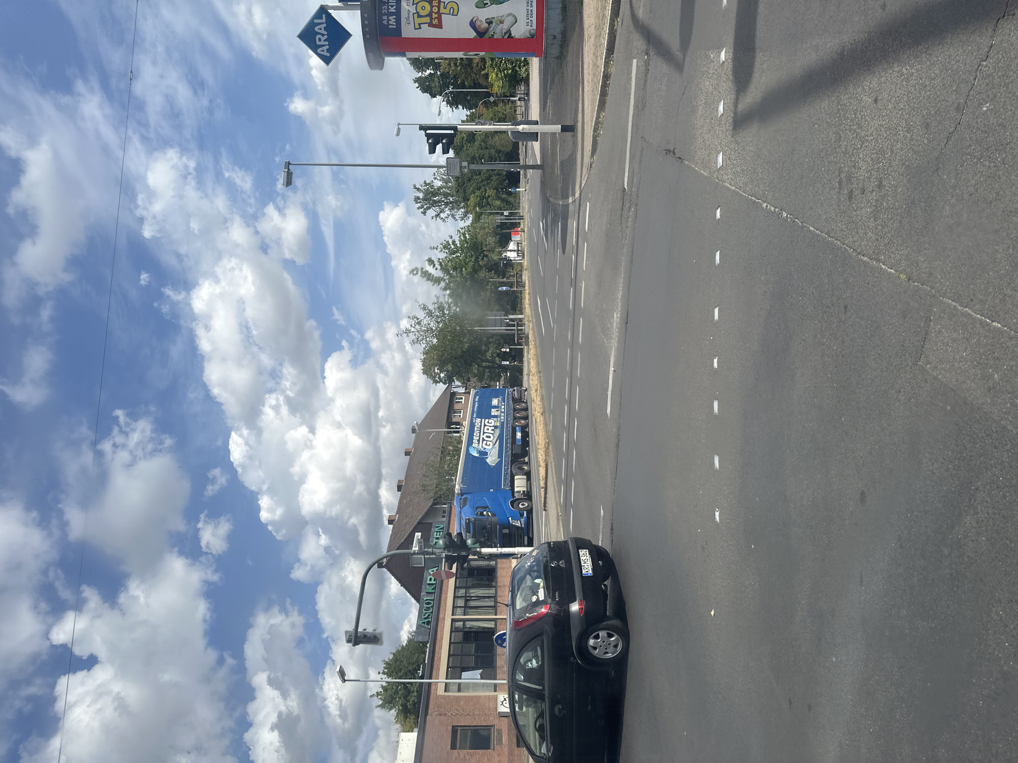 The image shows a street scene in Krefeld, Germany, on a cloudy day. A black Opel Corsa is stopped at a traffic light on the left side of the frame, with its license plate reading "KR MS 362". In the background, a large blue truck with "SPEDITION GORG" written on its side is passing through the intersection. To the left of the truck, a brick building with the sign "ASCOLIPA TEN" is visible. On the right side of the image, a billboard advertising "Disney PIXAR TOY STORY 5" is displayed. The sky is filled with white and gray clouds. The road is paved and shows lane markings. The overall environment appears to be an urban intersection with commercial and residential buildings.