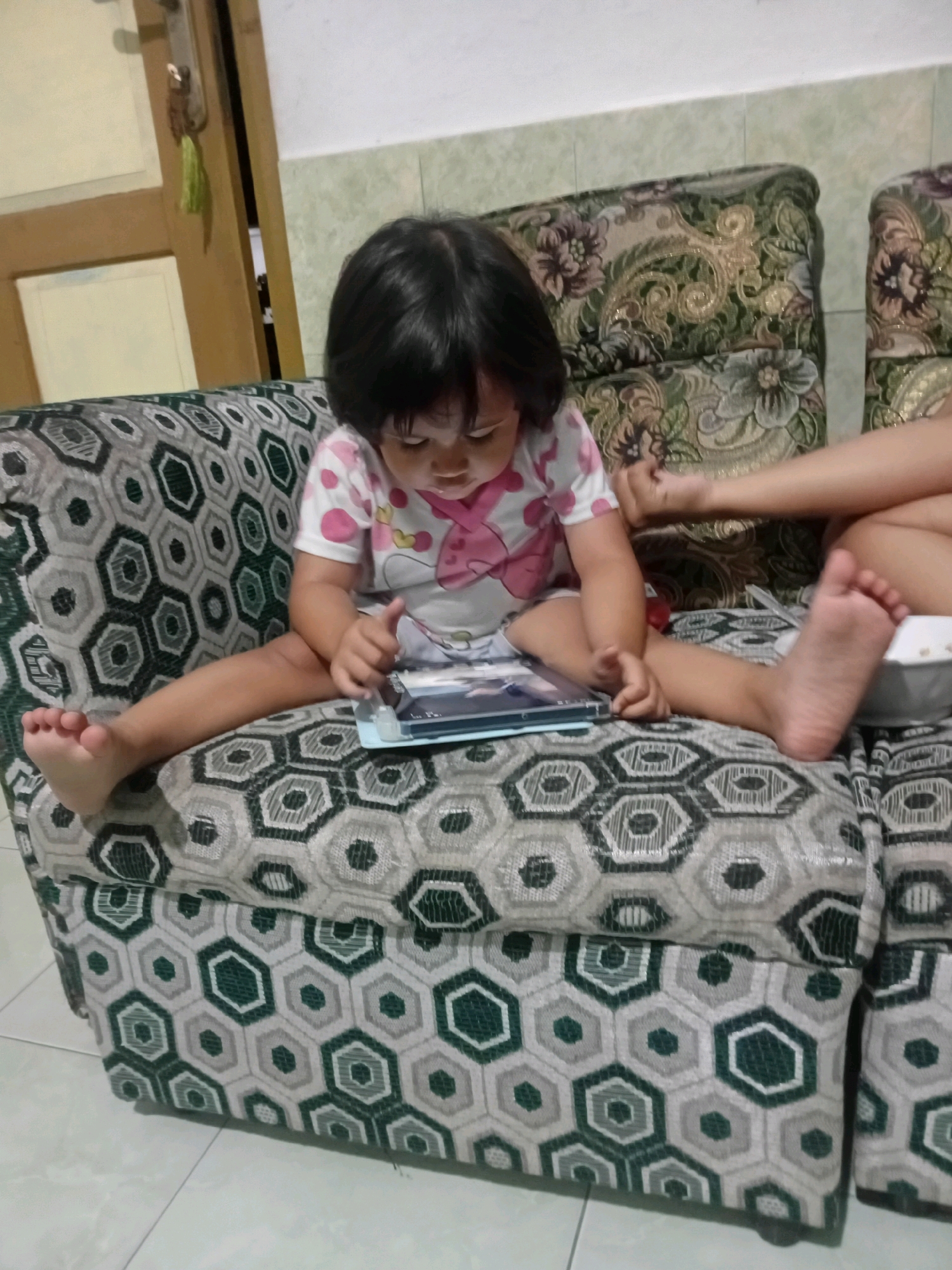 A young child with dark hair is sitting on a patterned sofa, looking at a tablet device. The child is wearing a white t-shirt with pink accents. Their legs are spread wide. Another person's arm is visible on the sofa to the right of the child. The sofa has a hexagonal pattern in shades of green and gray. In the background, a wooden door is slightly ajar, revealing a glimpse of a white wall. The floor appears to be tiled. This scene is taking place indoors, likely in a residential setting in Gebang, Indonesia.