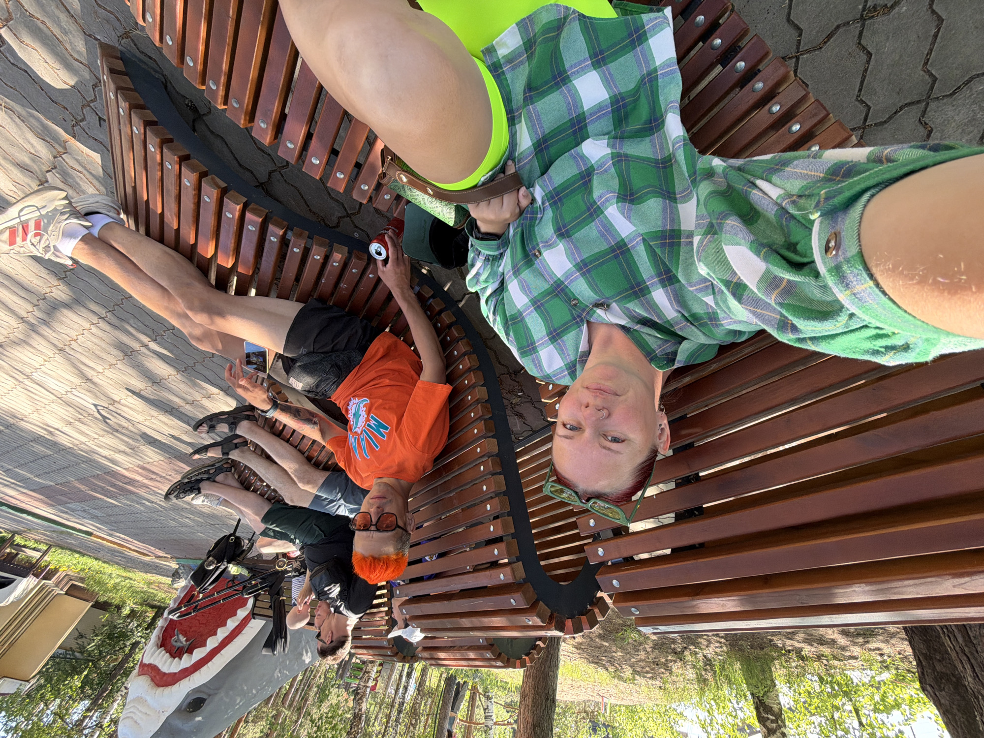 The image shows five people reclining on a wooden bench in what appears to be a park or public area. The photograph is taken from above, looking down at the group. The person closest to the camera is wearing a green and white plaid shirt and neon yellow shorts. Further down the bench is a person in an orange shirt with a logo, shorts, and orange hair and glasses. Beyond are three more people, one with dark hair and a backpack, and another holding a mobile phone. The bench is situated on a paved area next to grass and trees. In the background, there's a large shark sculpture. Based on the surroundings and shark sculpture, the location can be identified as Chaykovsky, Russia. The group appear to be relaxing on the bench.