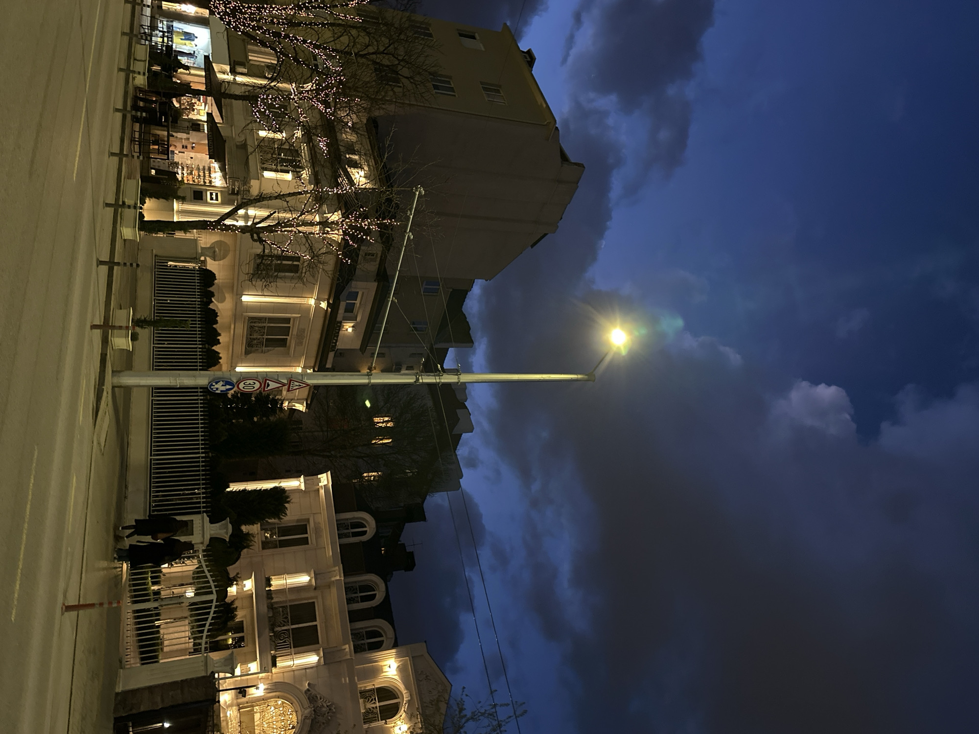 The image shows a street scene at night in Sofia, Bulgaria. The photo is taken from street level, looking towards a row of buildings. The buildings are illuminated with decorative lights. There are bare trees with string lights. A street lamp is visible in the middle of the image. The sky is dark with heavy clouds. There are a few people visible walking on the sidewalk. The road is wet. There are street signs near one of the buildings.