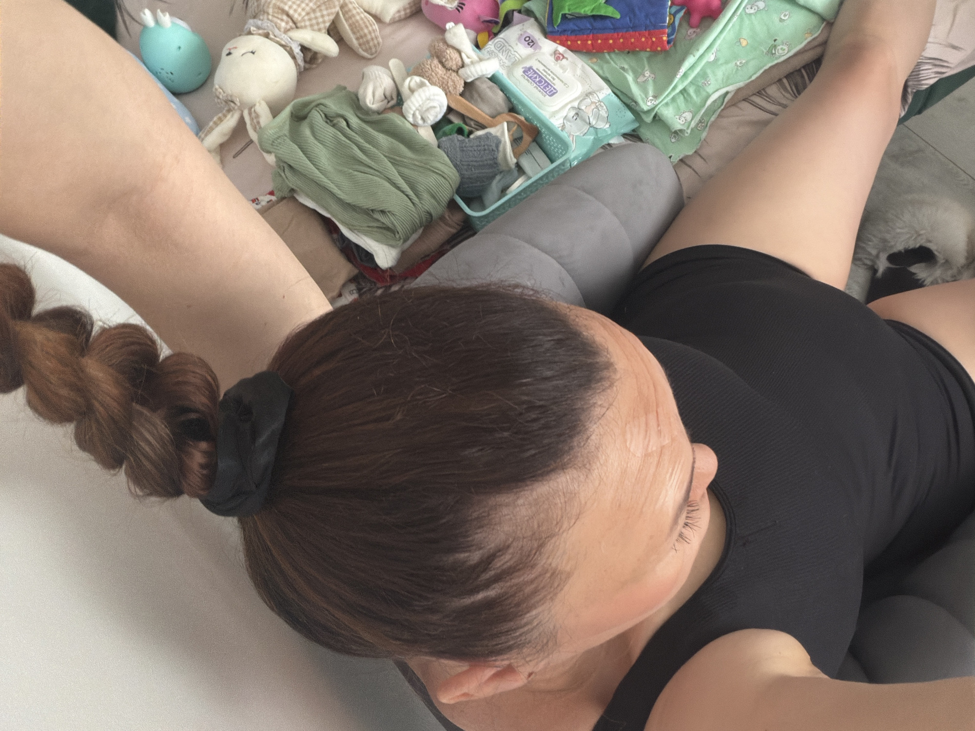 A high-angle shot reveals a woman with auburn hair styled in a braided ponytail secured with a black hair tie. She is wearing a black tank top. She is lying on a light-colored surface, possibly a bed or sofa. Around her are various items, including baby wipes, stuffed animals, baby clothes, and a gray pillow. The setting appears to be indoors, but specific details about the location in Chaikovsky, Russia, are not discernable from the image.