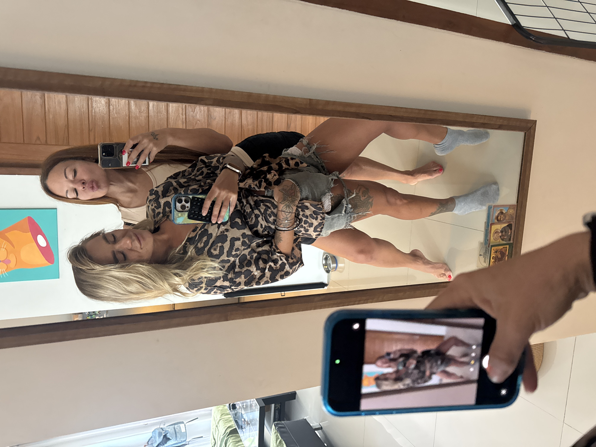 In Thep Krasatti, Thailand, two women are taking a selfie in front of a mirror. The woman on the left has blonde hair and is wearing a leopard-print shirt and ripped denim shorts. The woman on the right has long brown hair and is wearing a beige tank top and dark shorts. She is puckering her lips as if to kiss the camera. A third person's hand is visible in the foreground, holding a smartphone to capture the scene. The background shows a wall with wooden paneling and a framed picture of a cat. The floor is tiled.