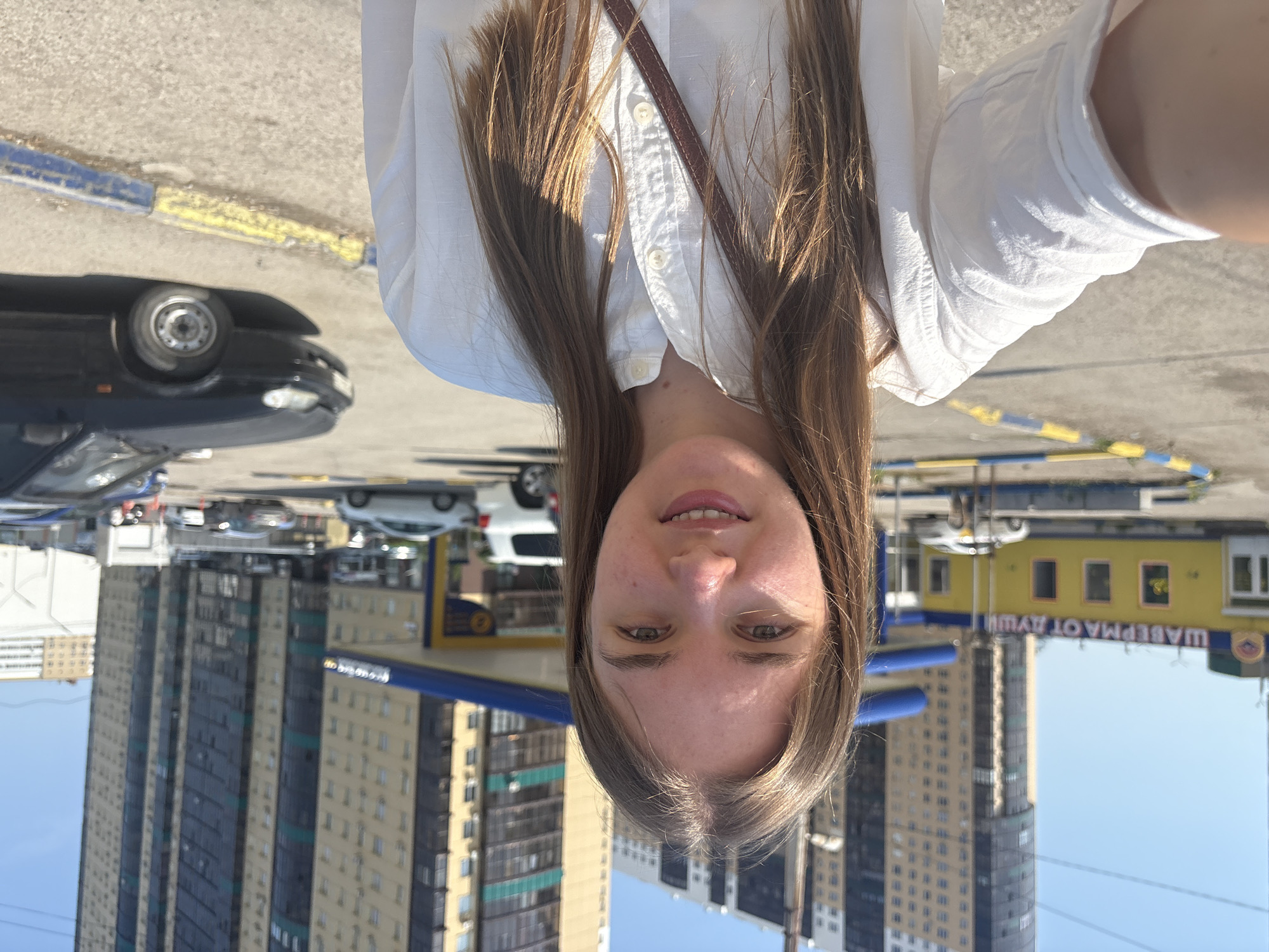 The image is a selfie of a woman with long, light brown hair, wearing a white collared shirt with a brown strap across her chest. The background features a cityscape, with tall buildings visible. There are also cars and what appears to be a small yellow building with Russian text, suggesting the location is likely in Russia. Based on the "SHAWERMA OT AYUU" (translated to Shawarma from Ayu) sign, the city is likely Perm, Russia. The sky is clear and sunny. The ground appears to be paved, possibly a parking lot or street.