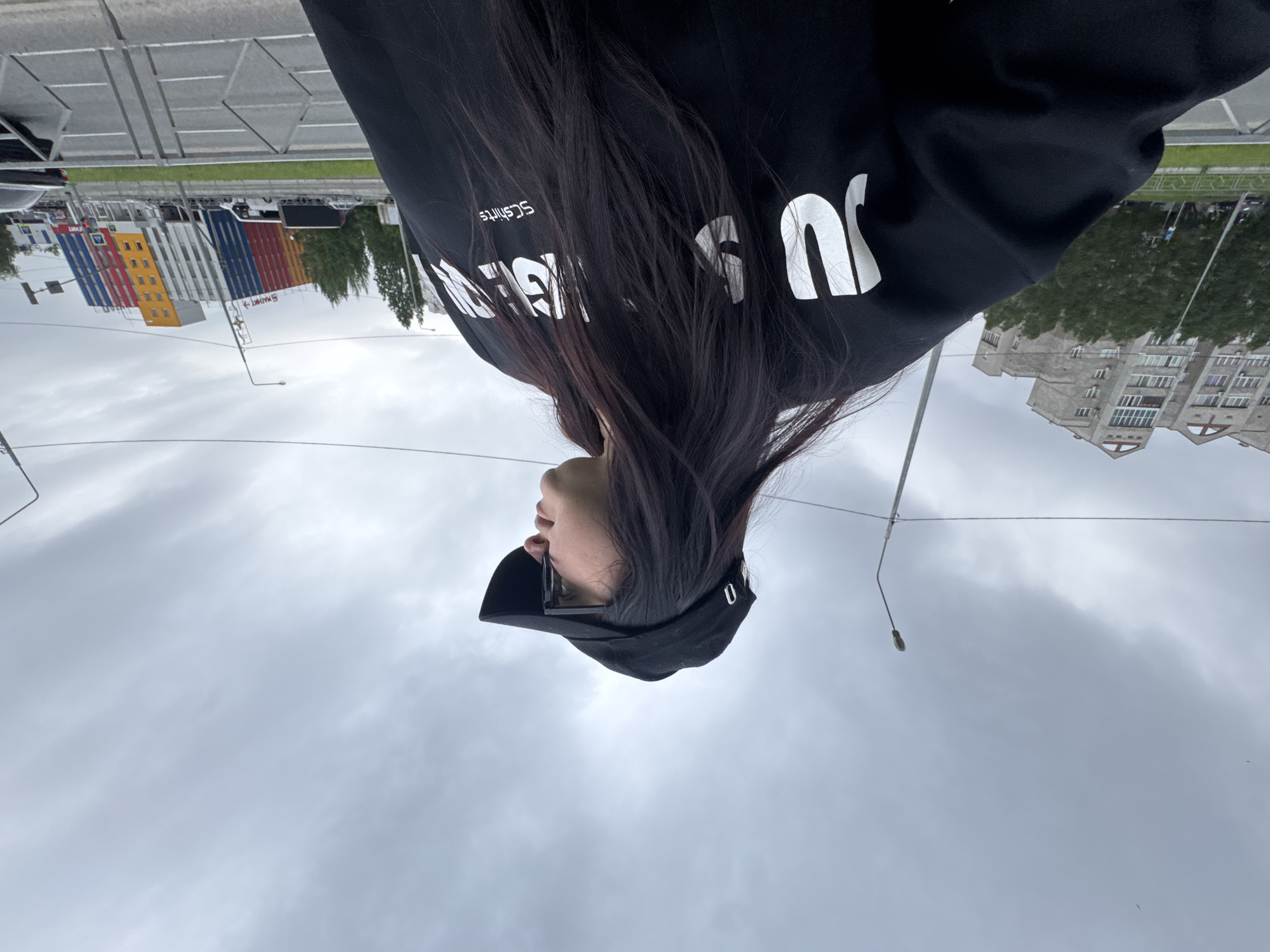 A medium shot depicts a woman wearing a black hoodie with white lettering, dark hair, and a black baseball cap. Her face is partially visible. The background features a city street, an apartment building, shipping containers in shades of red, blue, yellow, and white. A cloudy sky is overhead.