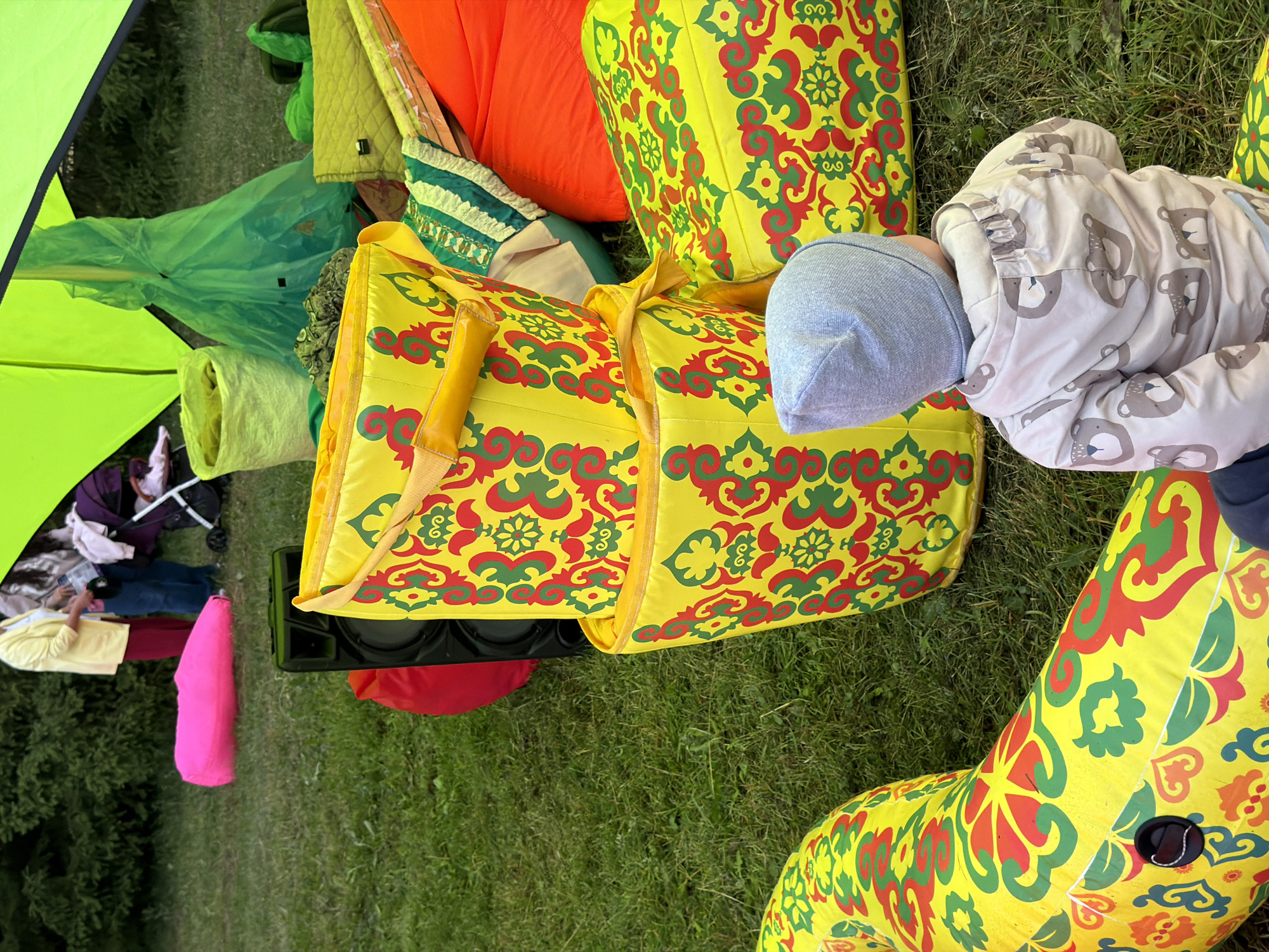 In an outdoor grassy area, an event appears to be set up with various colorful items. In the foreground, a young child wearing a light gray beanie and a beige jacket adorned with a pattern of teddy bear faces is facing away from the camera, seated on a large, yellow inflatable object decorated with red and green ornamental patterns. Behind the child, two stacked yellow cylindrical bags with similar red and green patterns are prominent. To the right of these bags, a bright orange beanbag chair and a lime green quilted bag are visible. Further back, a black speaker sits on the grass. In the background, several people are gathered, with a stroller and a bright pink inflatable object also present. Two large green tents or canopies are partially visible overhead. The ground is covered with green grass, and trees or shrubs form a backdrop.