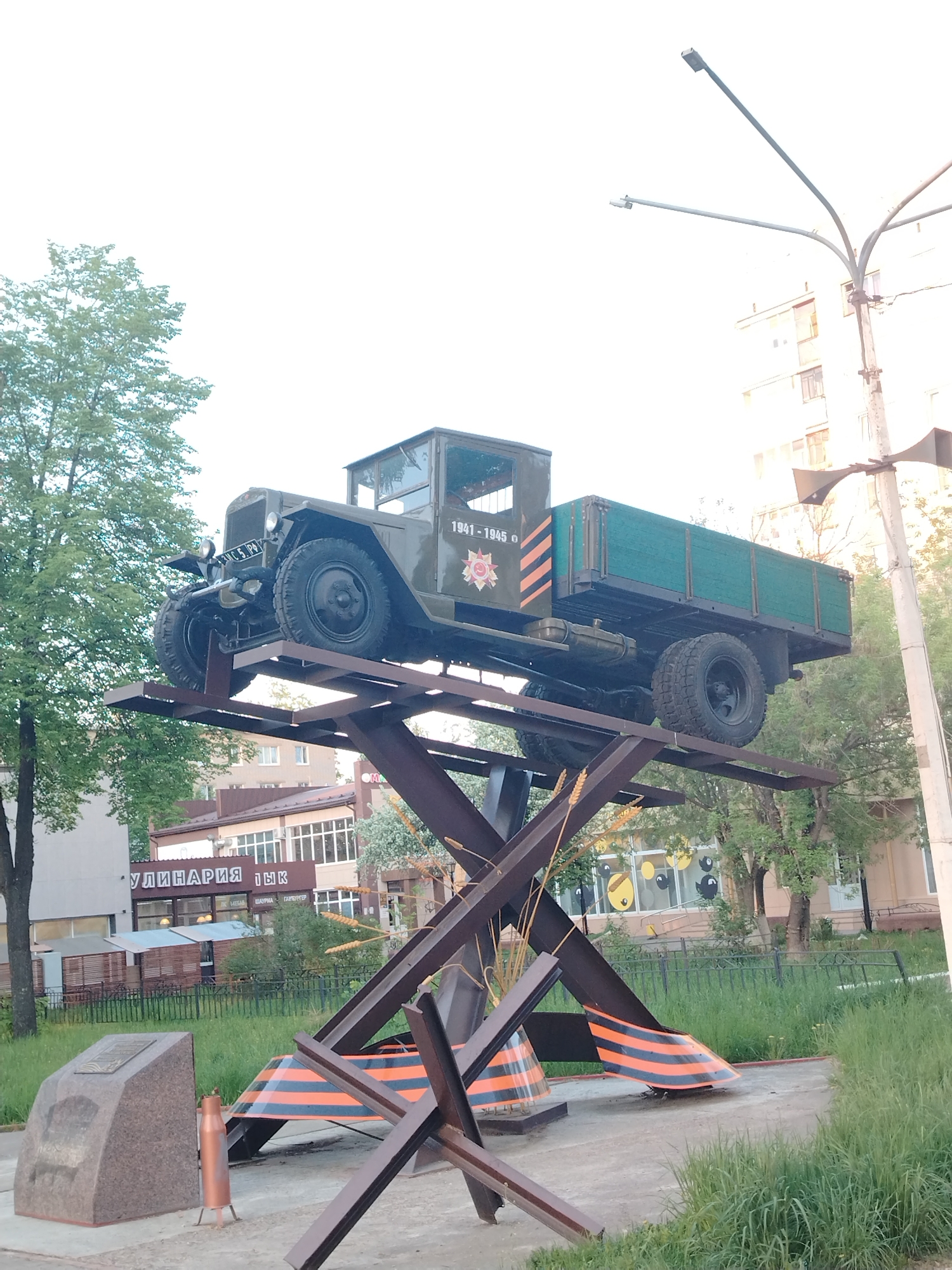 The image depicts a military truck monument located in the city of Chaikovsky, Russia. The truck, an older military model, is painted green with markings, is mounted on a dark brown metal structure resembling crossed anti-tank obstacles. The structure is further decorated with the orange and black striped "Ribbon of Saint George," a Russian military symbol. A small granite plaque stands nearby, presumably containing information about the monument. The background includes trees, grass, and buildings typical of an urban environment. A light post is also visible in the background.