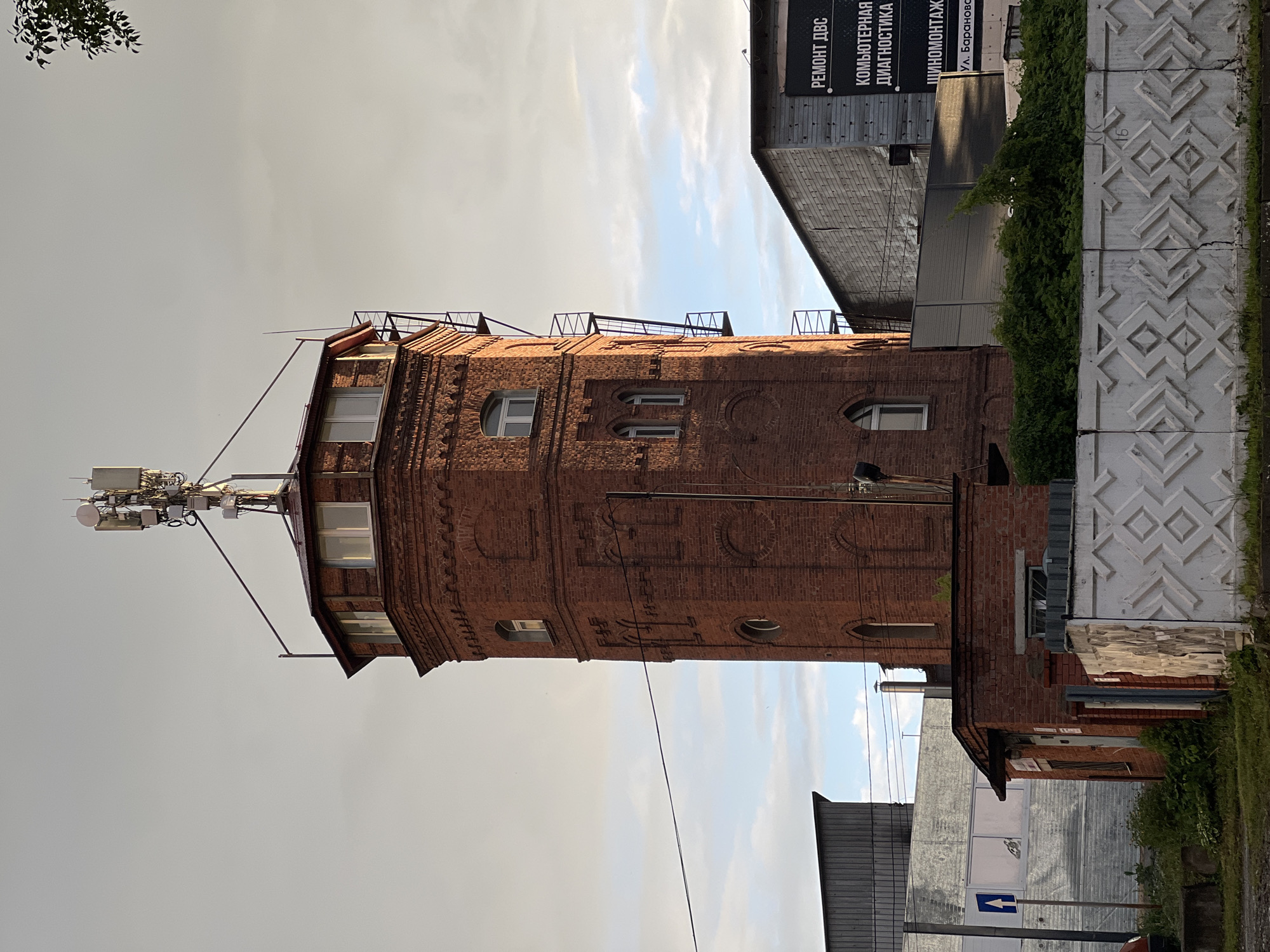 The image shows a tall, octagonal, red brick tower with a modern telecommunications antenna array at its summit. The tower has several windows of various shapes and sizes, and an external metal staircase is attached to its side. In front of the tower, there is a smaller red brick building with a corrugated metal roof. To the right of the tower, there is a large, grey, metal-clad building with a billboard displaying Russian text, including "РЕМОНТ ДВС" (engine repair) and "КОМПЬЮТЕРНАЯ ДИАГНОСТИКА" (computer diagnostics). A white, decorative concrete fence runs across the foreground. The sky is overcast with patches of blue sky visible. The location is indicated as Izhevsk, Russia. There are no people or animals visible in the image.