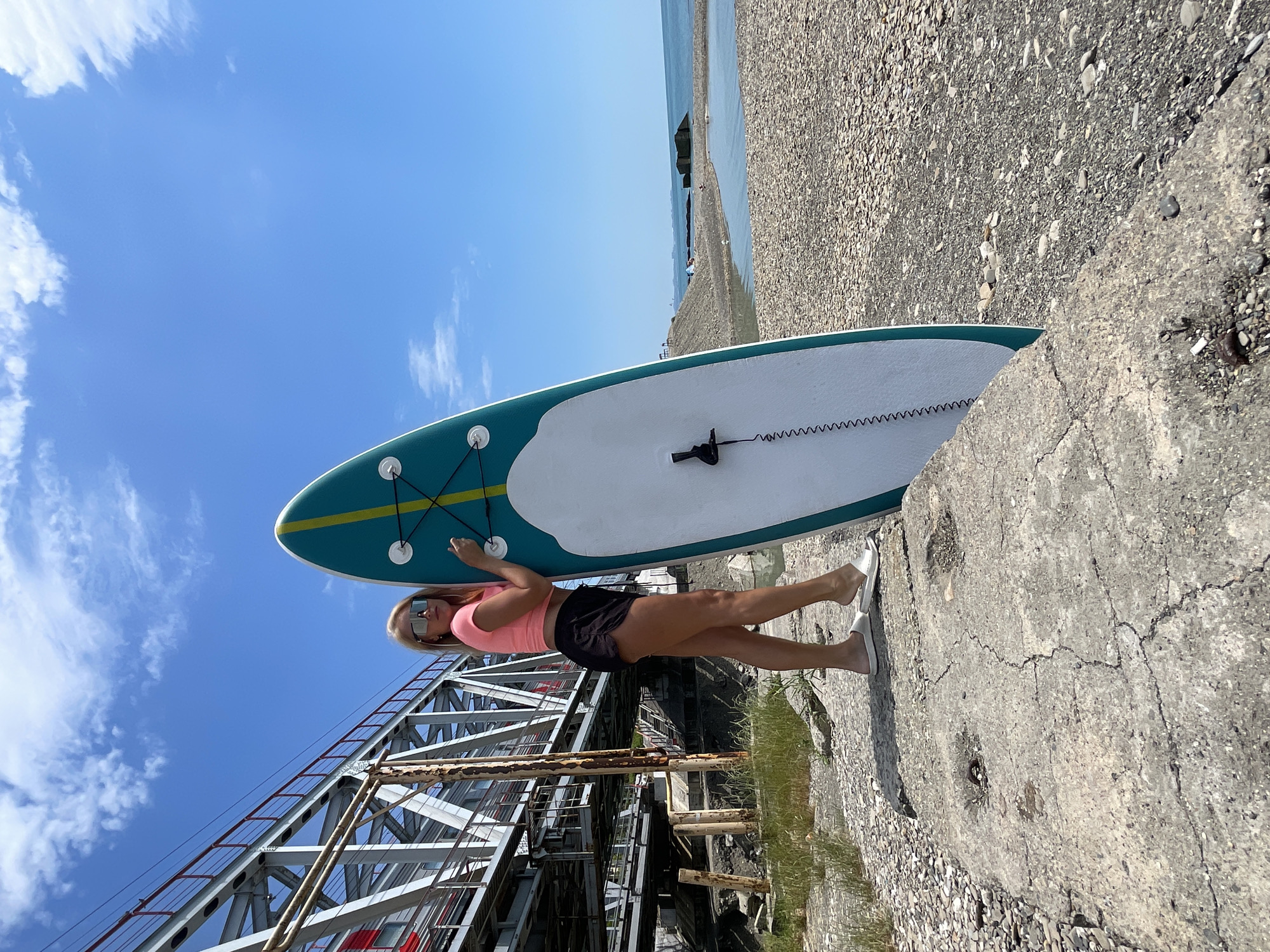 A blonde woman with sunglasses stands next to a teal and white paddleboard on a rocky shoreline, with a large metal bridge and a train in the background. The sky is blue with some clouds. The woman is wearing a pink crop top and black shorts. The paddleboard is upright, leaning against the woman's shoulder. The background shows a body of water, possibly a river or the sea, with a beach and some concrete structures. The ground is covered in pebbles and some dry grass.