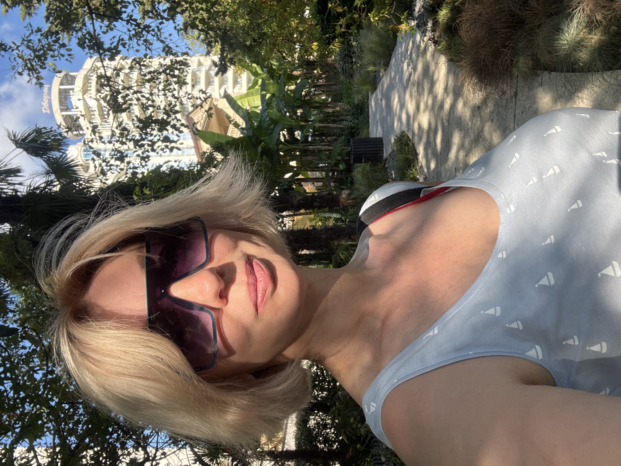 The image is a selfie taken by a woman with blonde hair and large sunglasses, wearing a light blue tank top with a sailboat pattern. She is outdoors in what appears to be a park or garden area. In the background, there is a tall, modern building with the Cyrillic word "Дворец" (Dvorets, meaning Palace) visible at the top. Lush green trees and plants surround the woman and the building. A paved walkway is visible, with shadows cast by the trees, suggesting it is a sunny day. The overall scene depicts a pleasant outdoor environment, likely in a resort or tourist area within Sochi, Russia.