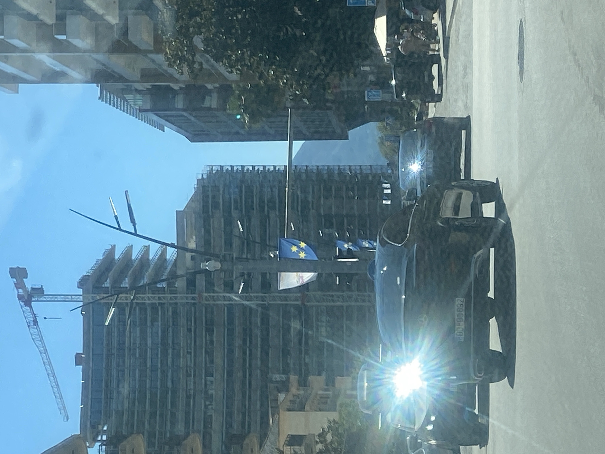 The image shows a street scene in Budva, Montenegro, viewed from inside a car. A construction crane is visible against a partly cloudy, blue sky. Several tall buildings, some under construction, line the street. European Union flags are on display. Two black cars are on the road. The license plate of the first car reads "BD-CG 892". Pedestrians are walking on the sidewalk.