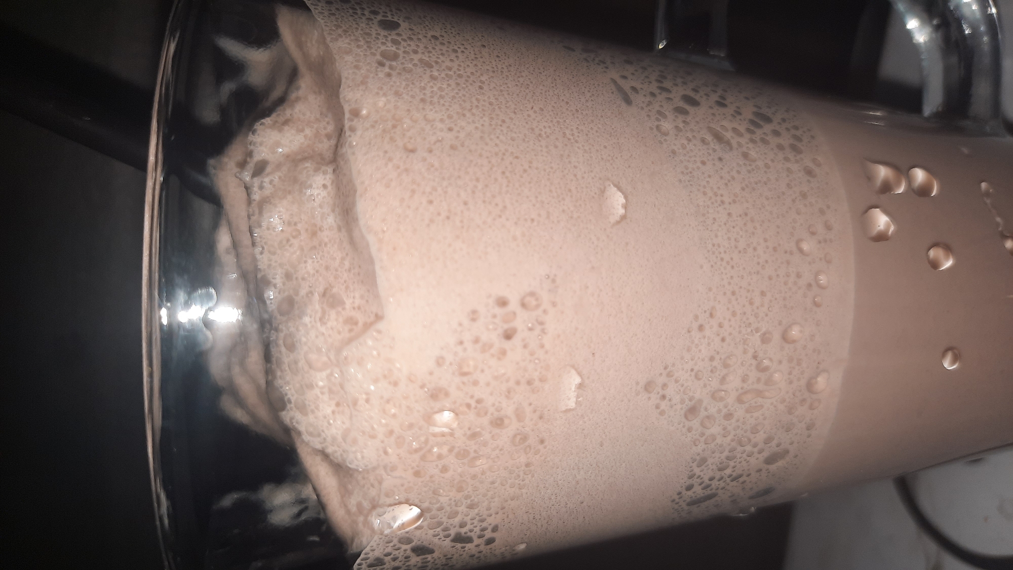A close-up shot shows a glass of chocolate milkshake. The drink is topped with a thick layer of froth, dotted with small bubbles. Water droplets are visible on the exterior of the glass, suggesting it is cold. The background is dark and out of focus. The image does not provide any information about the location in the city of Шклоў, Belarus.