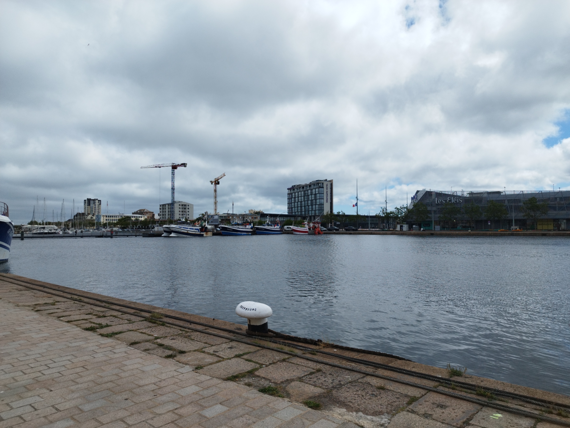 The image depicts a harbor scene in Cherbourg-en-Cotentin, France, on a cloudy day. In the foreground, a paved walkway made of rectangular stones runs along the edge of the water. A large white bollard with black trim, labeled "CHERBOURG", is situated on the walkway. Thick cables are laid on the stone surface. The water of the harbor is calm with small ripples.
Across the water, a line of docked boats is visible. These appear to be a mix of sailboats and motorboats, with masts of the sailboats extending upwards. Beyond the boats, a cityscape unfolds, characterized by several multi-story buildings. Prominent among these are two construction cranes, one blue and red and another yellow, indicating ongoing construction. One building prominently displays the name "Mercure Hotel". Another structure in the background has the signage "Les Eléis".
The sky is heavily overcast with large, grey clouds, though patches of lighter sky and possibly some blue are visible. A single bird is captured flying in the upper left quadrant of the sky. The overall scene suggests a functional port area with both maritime activity and urban development.