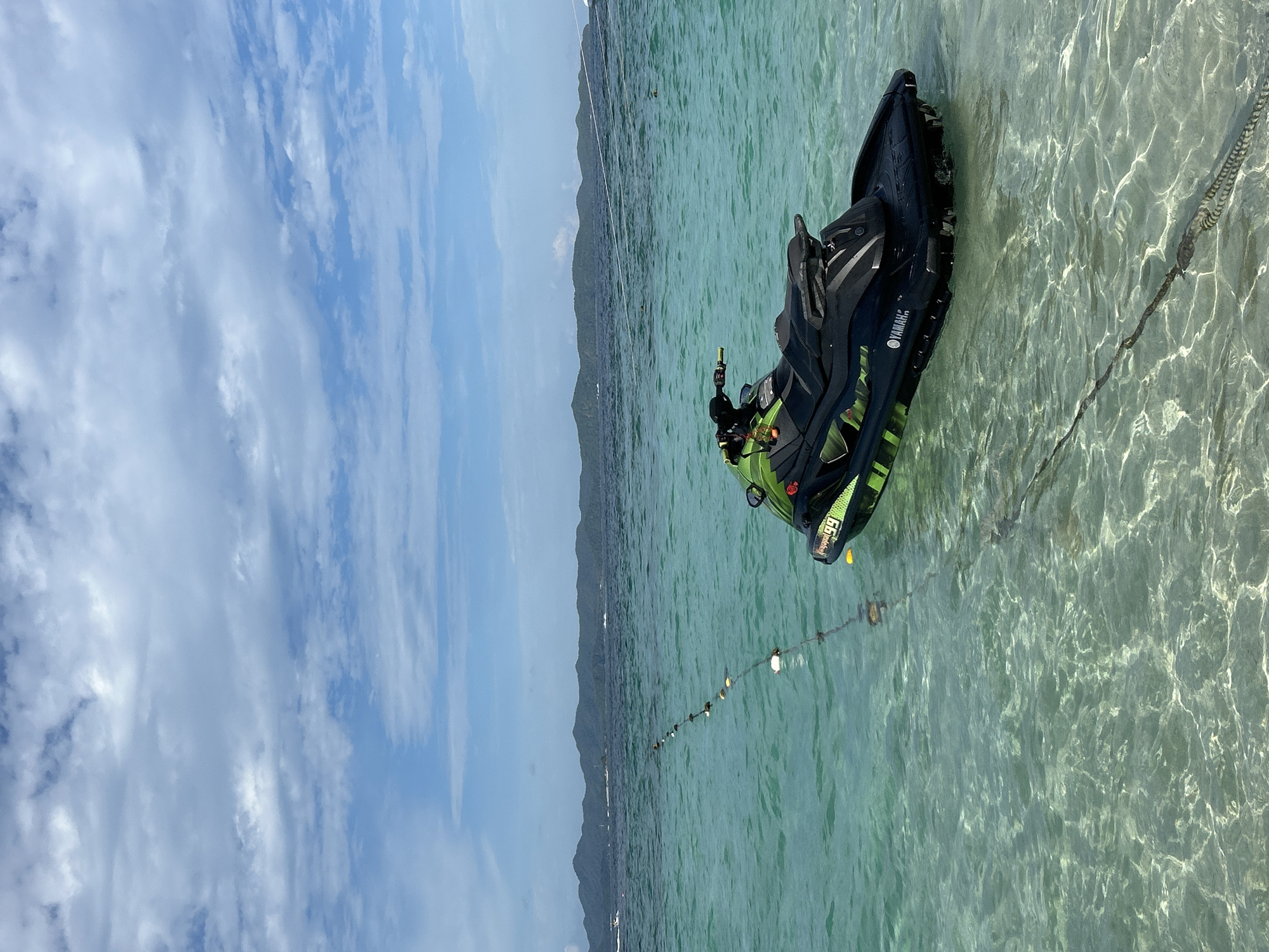A black and green jet ski, branded with "Yamaha" and the number "99" on its side, is docked in shallow, clear turquoise water. The jet ski is partially submerged, with its hull visible beneath the surface. A rope attached to the jet ski stretches towards the foreground, disappearing into the water. In the background, a line of buoys floats on the water, marking a boundary. Beyond the buoys, a distant, green, mountainous island range stretches across the horizon. The sky above is partly cloudy, with scattered white clouds against a blue backdrop. The overall scene depicts a serene coastal environment.
