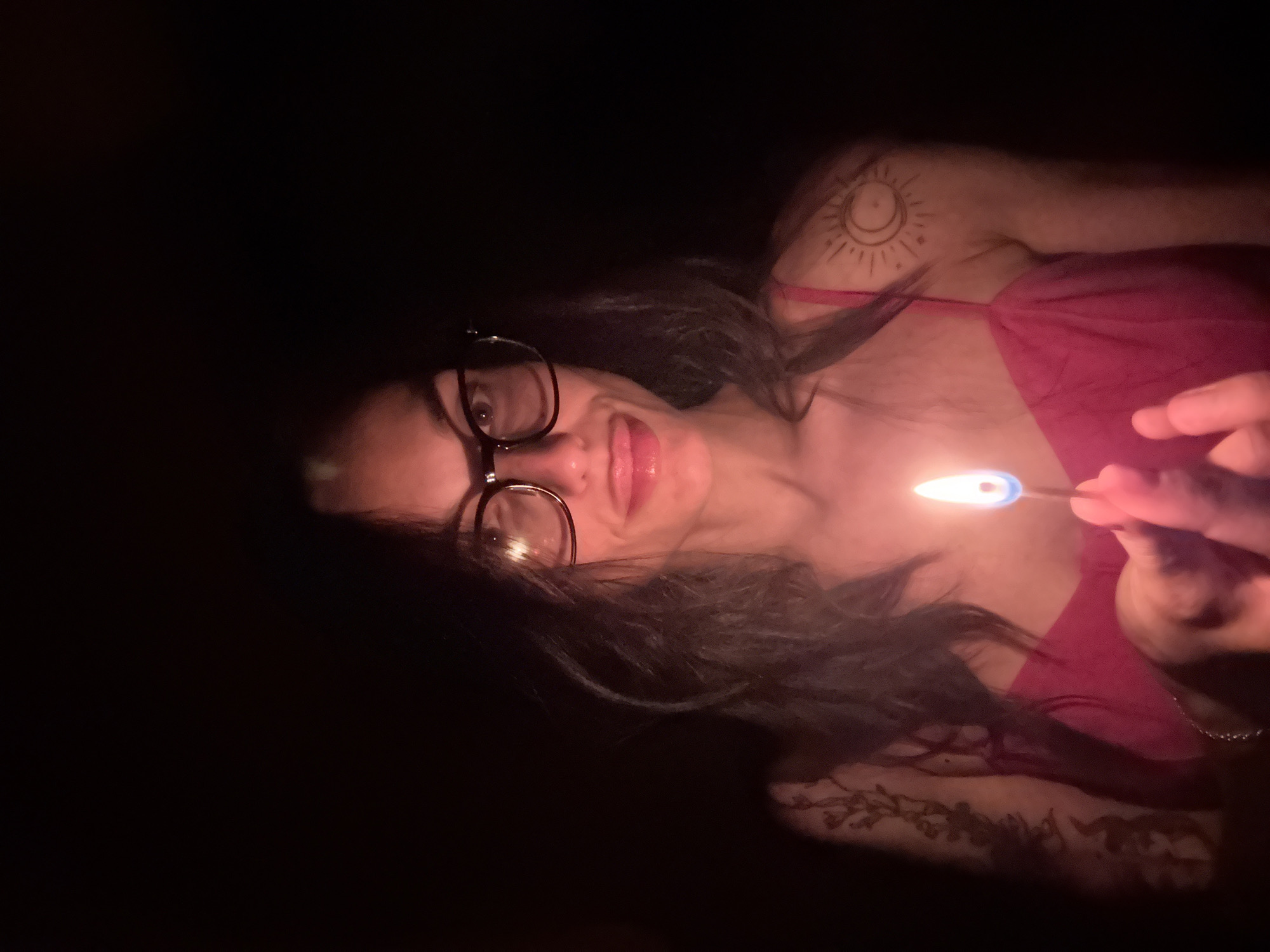 The image, taken in Pleven, Bulgaria, captures a female-presenting individual in a very dark setting, likely at night or in a dimly lit room. The subject is shown at an angle, as if lying down, with her head oriented towards the left side of the frame. She wears dark-rimmed glasses and has long dark hair. Her expression is a playful smile or smirk, with her tongue slightly extended.
She is wearing a pink or magenta-colored top with thin straps. Her right hand holds a lit match, which acts as the sole light source, casting a warm, glowing light on her face and upper body. The match flame exhibits a distinct blue at its base transitioning to orange at the top.
Notable details include a tattoo on her right shoulder/upper arm, depicting a stylized sun and crescent moon. Another tattoo with a detailed, possibly floral or leafy, pattern is visible on her lower left arm. The background is completely black, creating a stark contrast with the illuminated subject. The overall mood is intimate and somewhat mysterious due to the selective lighting from the match. No visible text is present in the image.