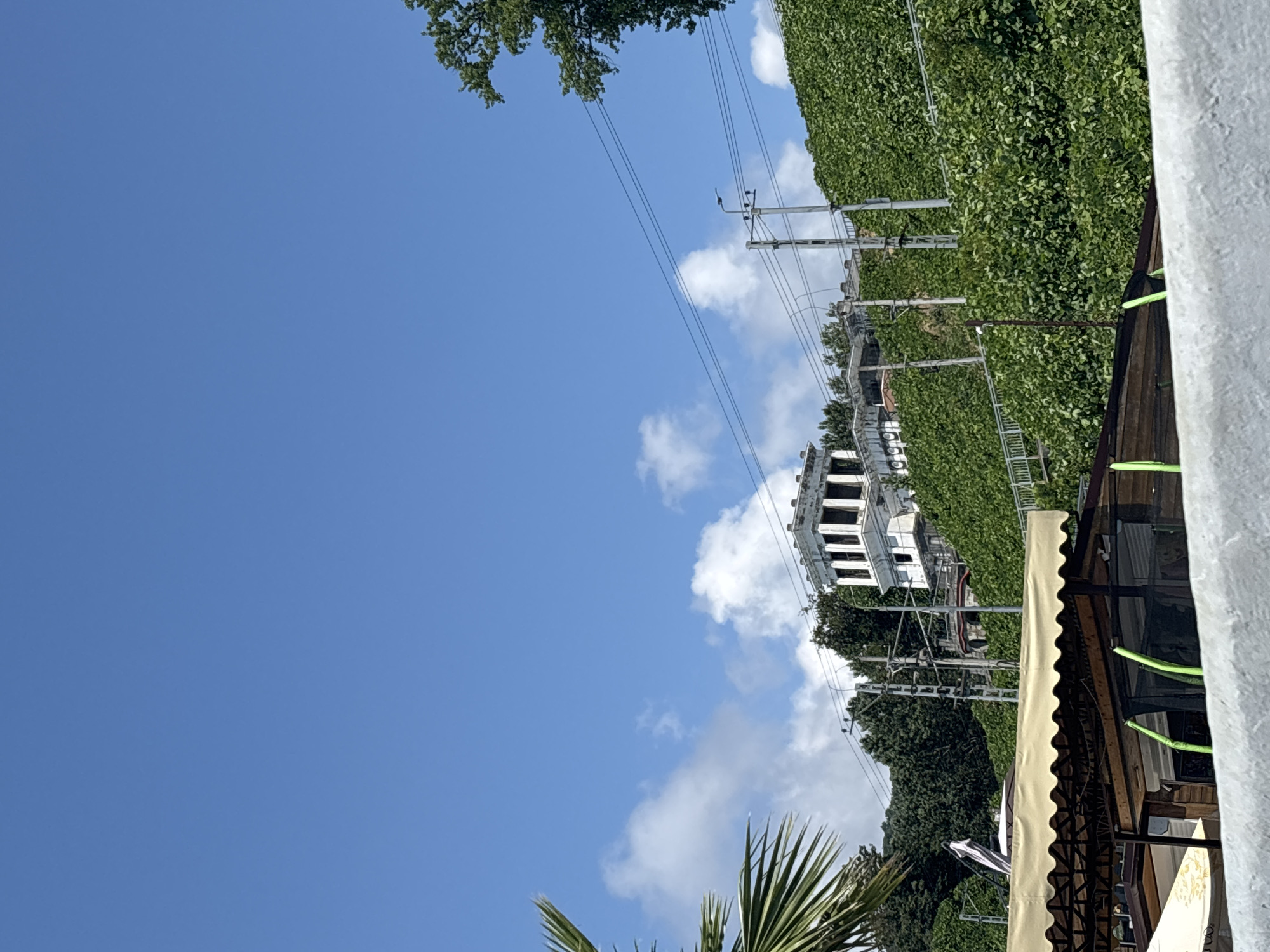 The image depicts a sunny day in Sochi, Russia. In the foreground, a beige awning covers a section of an outdoor establishment, possibly a cafe or market. The awning is supported by a dark metal frame. Behind this, greenery covers a steep hillside, with a white building featuring multiple columns and rectangular openings visible on its upper portion. Electrical poles and wires are also present along the slope, interspersed with the vegetation. The sky above is a vibrant blue with scattered white clouds. A palm frond is visible in the lower left corner.