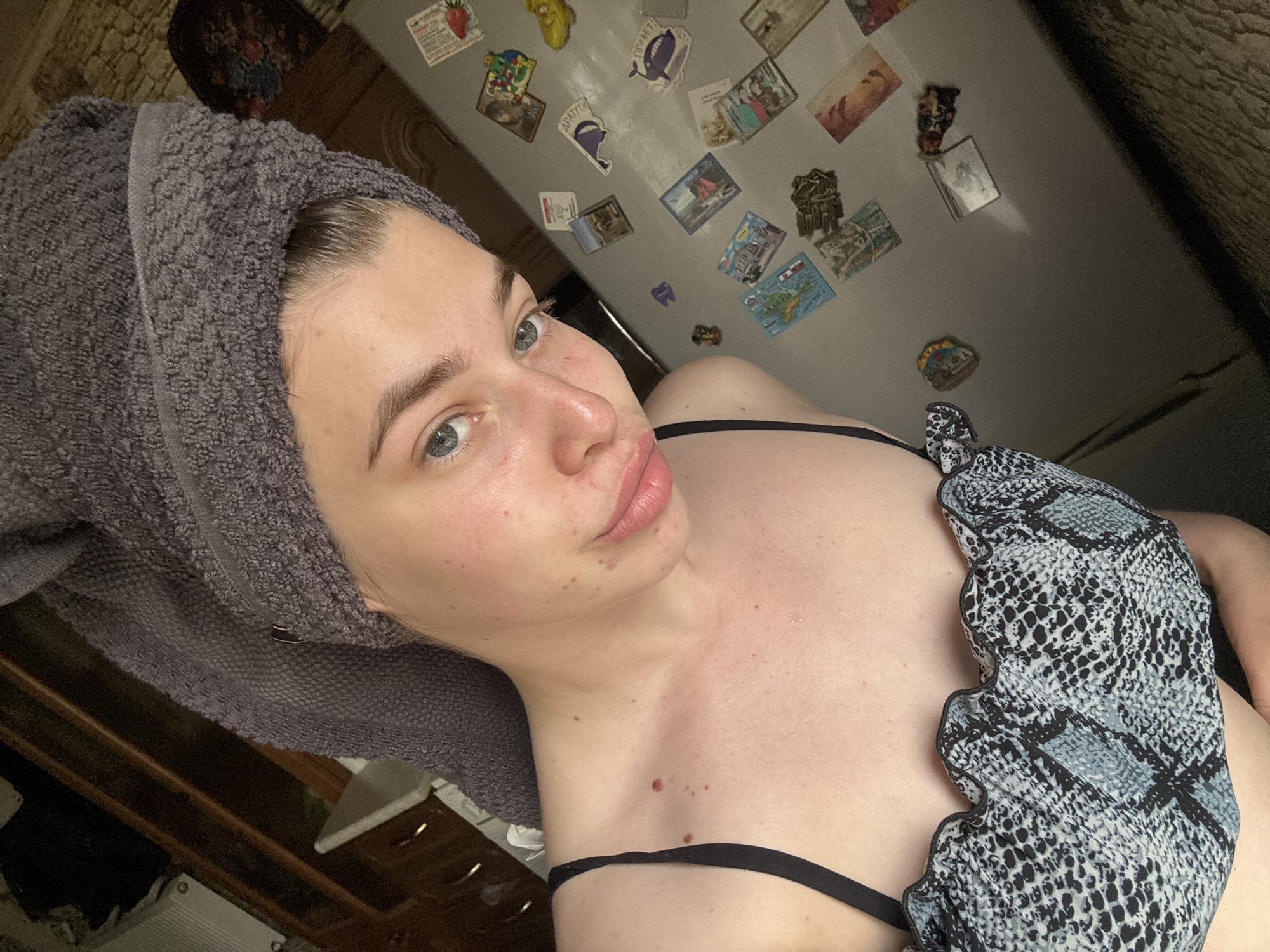 A selfie of a fair-skinned woman with a towel wrapped around her head. She is wearing a black and grey patterned top. The background includes a refrigerator covered with magnets and a textured wall. The scene appears to be indoors, possibly in a domestic setting. There is no evidence in this image to suggest a specific location in the city of Мещёры, Russia.