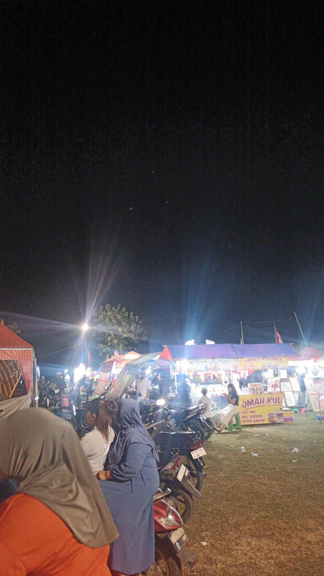 The image depicts a nighttime outdoor scene, likely a market or fair, in Ketemasdungus, Indonesia. The foreground is dominated by three individuals, two women and a person in an orange garment with a grey head covering. One woman, wearing a blue hijab and blue clothing, is seated on a motorcycle. Another woman, in a white top, is seated behind her on the same motorcycle. The third person, partially visible, is wearing an orange shirt and a grey head covering.
Behind them, the scene is illuminated by various lights from stalls and streetlights. A large tree is visible in the mid-ground, with light beams radiating from a streetlamp. Several stalls are set up, with visible signage including "OMAH KULINER" and "POP ICE". Motorcycles are parked in front of the stalls. The ground appears to be covered in dry grass or dirt. The sky is dark, indicating it is nighttime, with some lens flares visible from the lights.