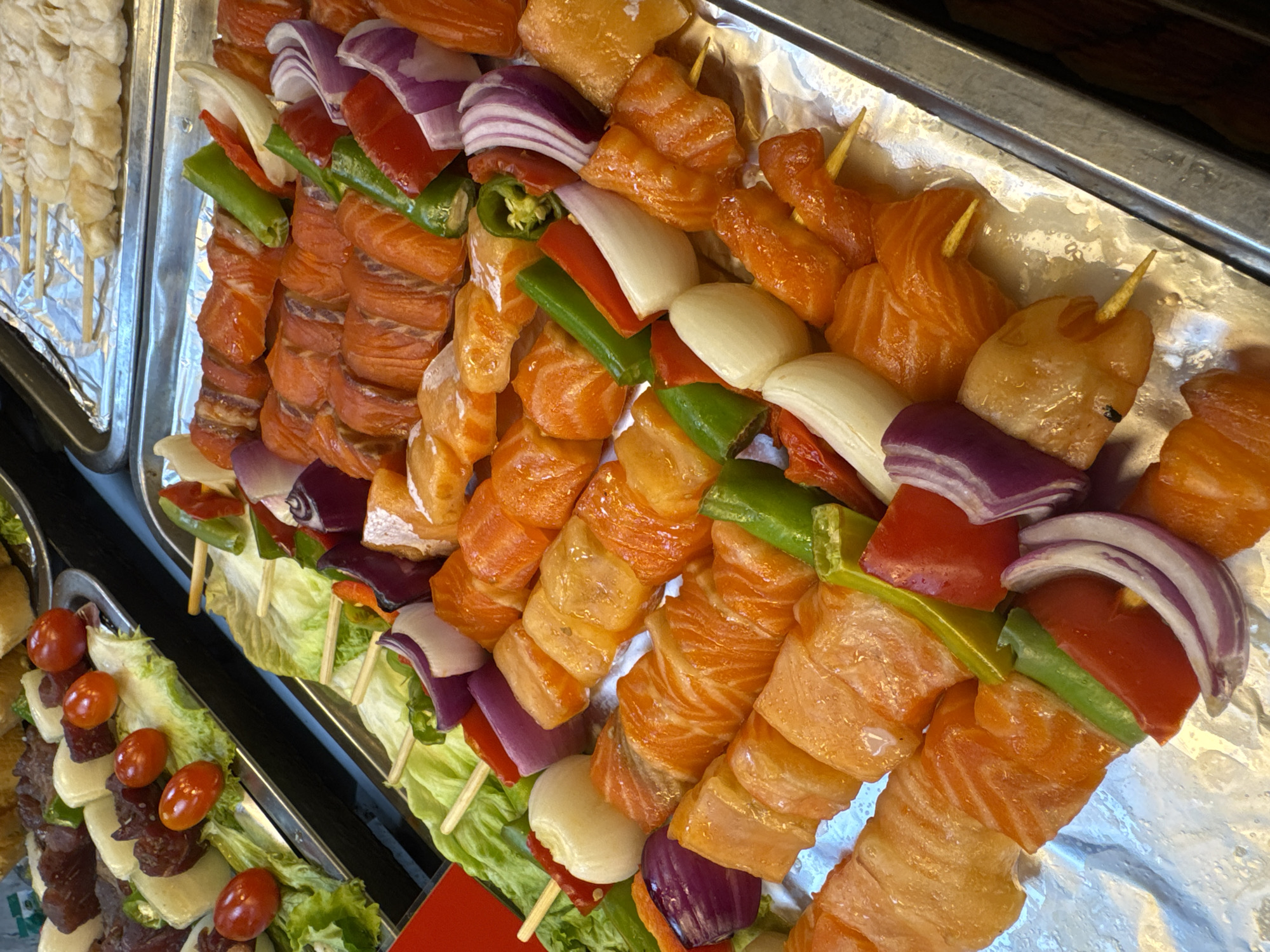 A high-angle shot displays skewers of salmon and vegetables on aluminum trays in Pattaya City, Thailand. The skewers feature chunks of salmon alternating with red onion slices, green bell pepper pieces, red bell pepper chunks, and garlic cloves. Some skewers rest on a bed of lettuce. Another tray contains white skewers. A tray beside these holds skewers of meat, cheese, and tomatoes on lettuce.