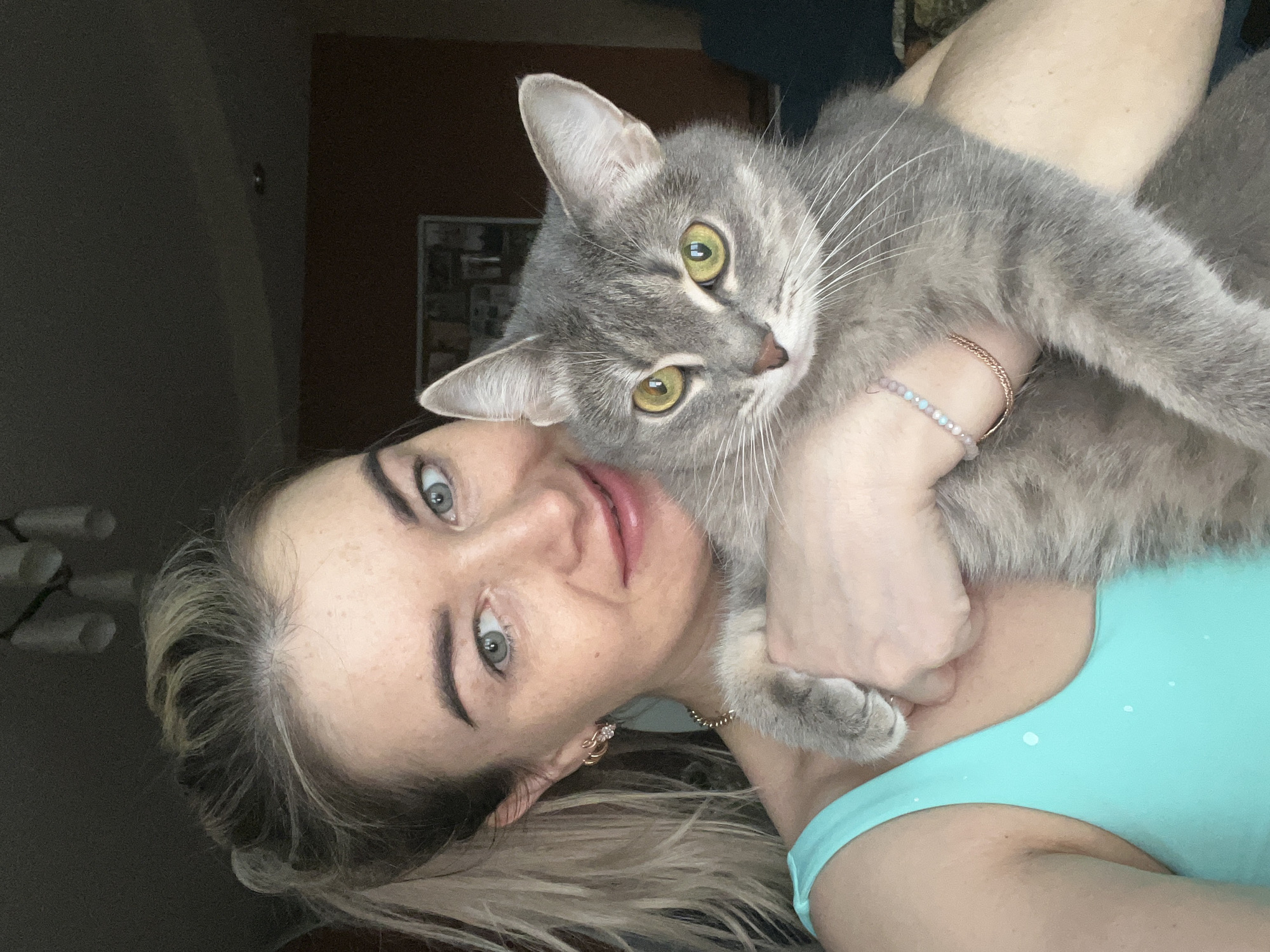 A woman with blonde hair and blue eyes is holding a grey tabby cat. The woman is wearing a light blue tank top. The cat has green eyes. The background is blurry and dark, with a door visible. It is not possible to confirm that this image was taken in Chaikovsky, Russia, from this image alone.