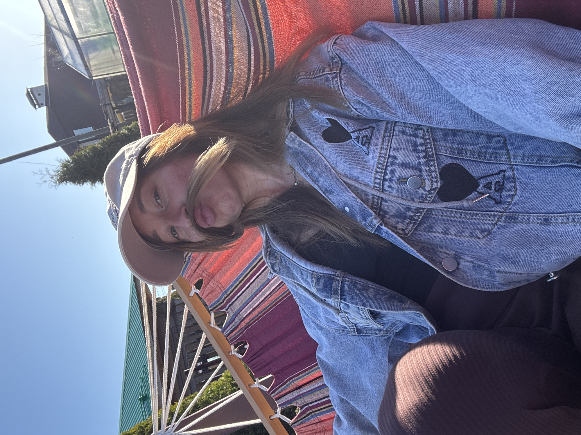 A woman is lying in a hammock outdoors. She is wearing a tan baseball cap, a blue denim jacket with a black heart patch, and a black top. The hammock has red, orange, and blue stripes. In the background, there is a blue sky, green trees, and a building. The location is in the city of Прикамский, Russia.