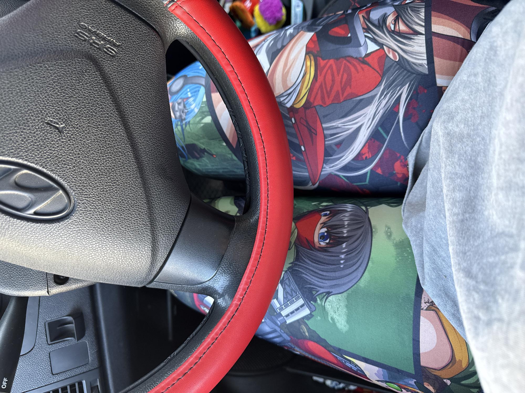 The image provides a close-up view of a personalized car interior, captured from the driver's perspective looking towards the passenger side, set in Chaikovsky, Russia. The foreground is dominated by a black plastic steering wheel, partially visible, which has a vibrant red faux leather cover on its rim, accentuated by black stitching. The Hyundai logo is visible on the lower spoke of the steering wheel, and the text "SRS Airbag" is embossed on the airbag cover, accompanied by a small horn symbol. Below the steering wheel, parts of the dashboard and a lever with the text "OFF" are visible.
On the passenger seat and dashboard, several anime-style body pillows (dakimakura) are prominently displayed. The pillow closest to the viewer features a character with dark hair, blue eyes, and a red mask, holding what appears to be a firearm, set against a green, patterned background. Another pillow, positioned behind and slightly above the first, depicts a character with long, flowing white hair, dressed in red and dark attire, with a serious expression. A third, less distinct pillow in the background shows a blue-haired character wearing goggles. A light-colored fabric, possibly a blanket or clothing, partially covers some of the pillows. In the blurred background, hints of bright yellow and purple objects suggest other items are present in the car.
The car interior is well-lit by natural daylight, indicating it is daytime. The vehicle appears to be stationary, highlighting the owner's distinctive customization reflecting their interest in anime culture.