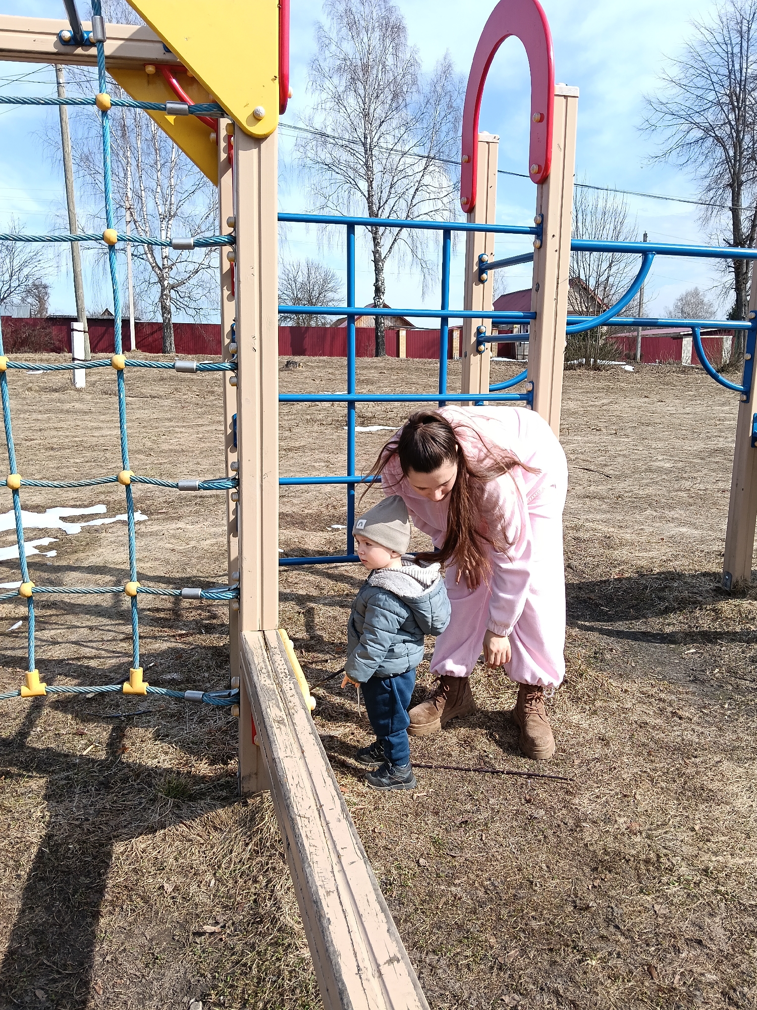 In Красный Маяк, Russia, a young child in a grey jacket, blue pants, and dark shoes stands on dry grass near a playground structure. A woman in a pink jumpsuit and brown boots bends over, seemingly interacting with the child. The playground features blue climbing bars, a rope net with yellow connectors, and beige wood supports. The ground is covered in dry grass and patches of snow. In the background, bare trees are visible under a blue sky. A red fence lines the perimeter.