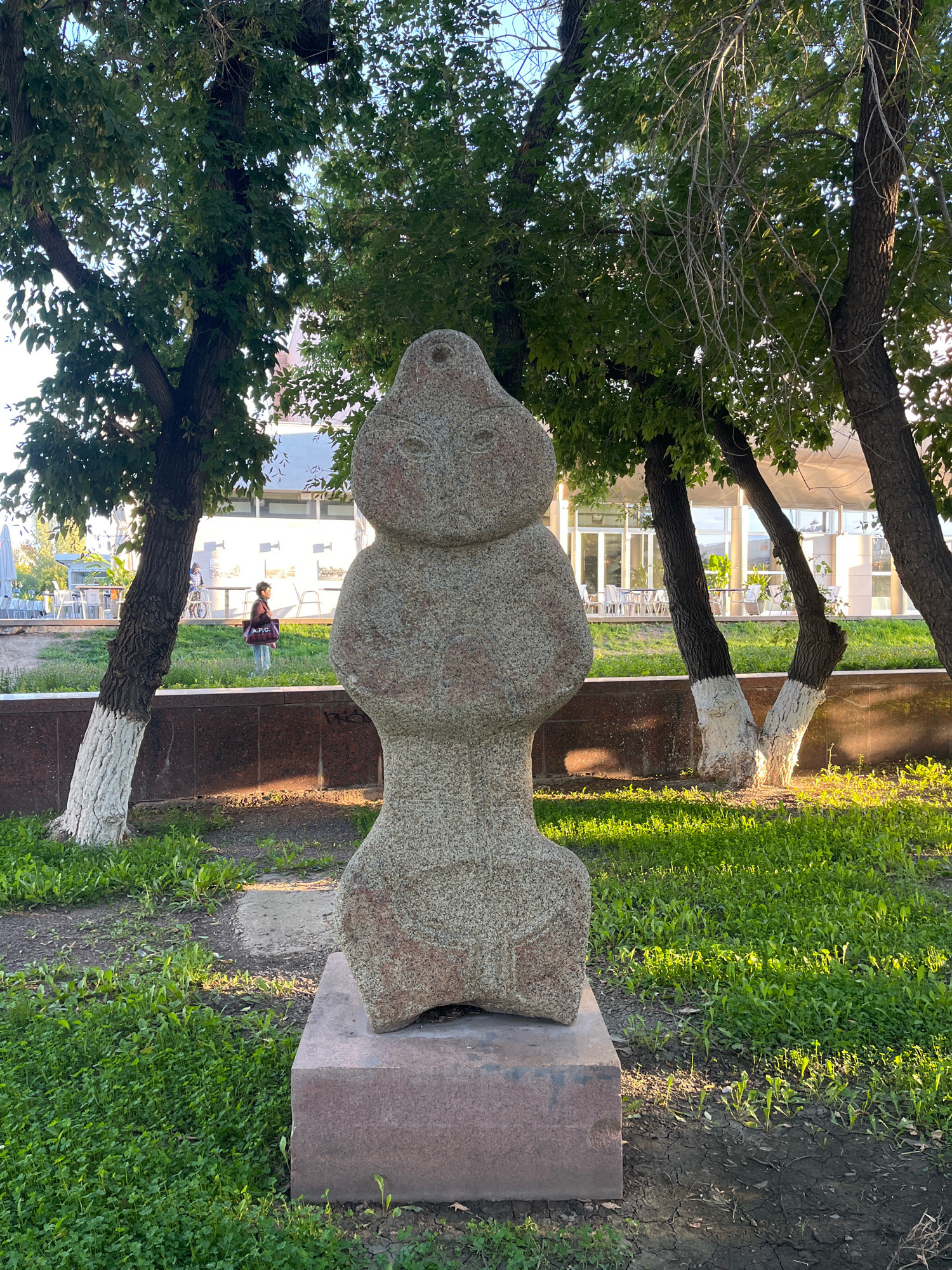 In Astana, Kazakhstan, a stone statue stands in a park-like setting. The statue, a monolithic representation of a human figure with stylized features, is positioned on a rectangular pink granite pedestal. It is made of rough-hewn stone with visible texture. The statue is centrally located in the foreground, with green grass and some dirt patches surrounding its base.
Behind the statue, a dark brown marble retaining wall is visible. Beyond the wall, a grassy area slopes upwards, leading to a building with large glass windows and white trim. Several trees with green leaves and light-colored, whitewashed trunks are scattered throughout the scene, casting shadows on the ground. Some of the trees have bare branches reaching upwards.
In the middle ground, a person is walking or standing on the grass, carrying a dark bag with a visible red logo. This person is viewed from the side, facing away from the statue. In the background, outdoor seating areas with tables and chairs are visible near the building, suggesting a cafe or restaurant. The lighting suggests it is daytime, with the sun casting distinct shadows and highlighting the textures of the stone and grass. The overall impression is of a public outdoor space, likely a park or a square within the city of Astana.