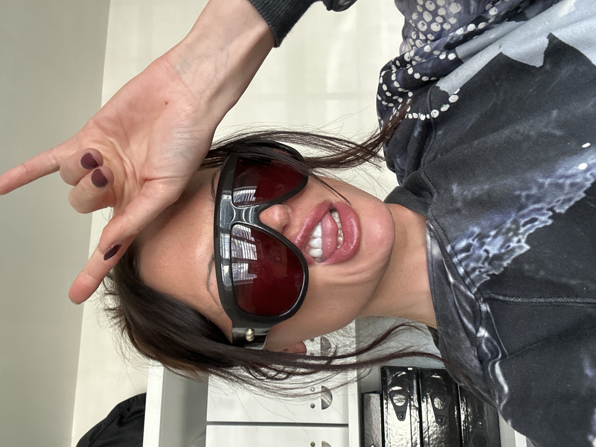 The image displays a headshot of a person, rotated 90 degrees clockwise. The individual is making a "rock on" hand gesture with their left hand, with the index and pinky fingers extended and the thumb holding down the middle and ring fingers. The fingernails are painted with a dark burgundy or plum-colored polish.
The person is wearing large, black-framed sunglasses with dark red-tinted lenses, obscuring their eyes. Their mouth is open, with the tongue partially visible and slightly curled. Dark brown hair frames the face, with some strands falling across the forehead and sunglasses.
The individual is dressed in a dark-colored hoodie or sweatshirt. The garment features a black or dark grey base with lighter, abstract, cloud-like patterns. A portion of the clothing, possibly a hood or collar, displays a contrasting pattern of white polka dots on a darker fabric.
The background consists of a plain, light-colored wall, suggesting an indoor setting. Towards the bottom right of the image (as it is oriented), several shiny, dark binders or folders with metallic clasps are visible, resting on a white surface, further indicating an indoor environment like an office or study. The overall impression conveyed by the pose and accessories is one of playfulness, confidence, or a rebellious attitude. No text is visible within the image.