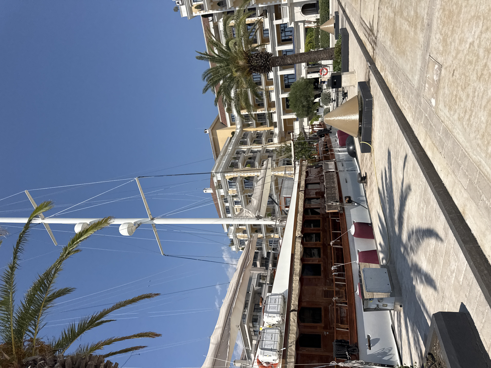 The image depicts a sunlit scene in Tivat, Montenegro, likely within the Porto Montenegro marina. Dominating the foreground is a large, classic sailing yacht with a white hull and prominent wooden trim, docked alongside a paved promenade. The yacht features several white life preservers and large, rolled-up sails. Behind the yacht, a series of modern, multi-story buildings with cream-colored facades, balconies, and red-tiled roofs are visible, indicating a resort or residential complex. Two large palm trees frame the scene, one on the left and another on the right, with their fronds casting shadows on the walkway. The promenade is paved with light-colored stones and features dark gray curbing. Several decorative conical sculptures, possibly fountains, are placed along the promenade. The sky is a clear, vibrant blue. No people are visible in the frame.