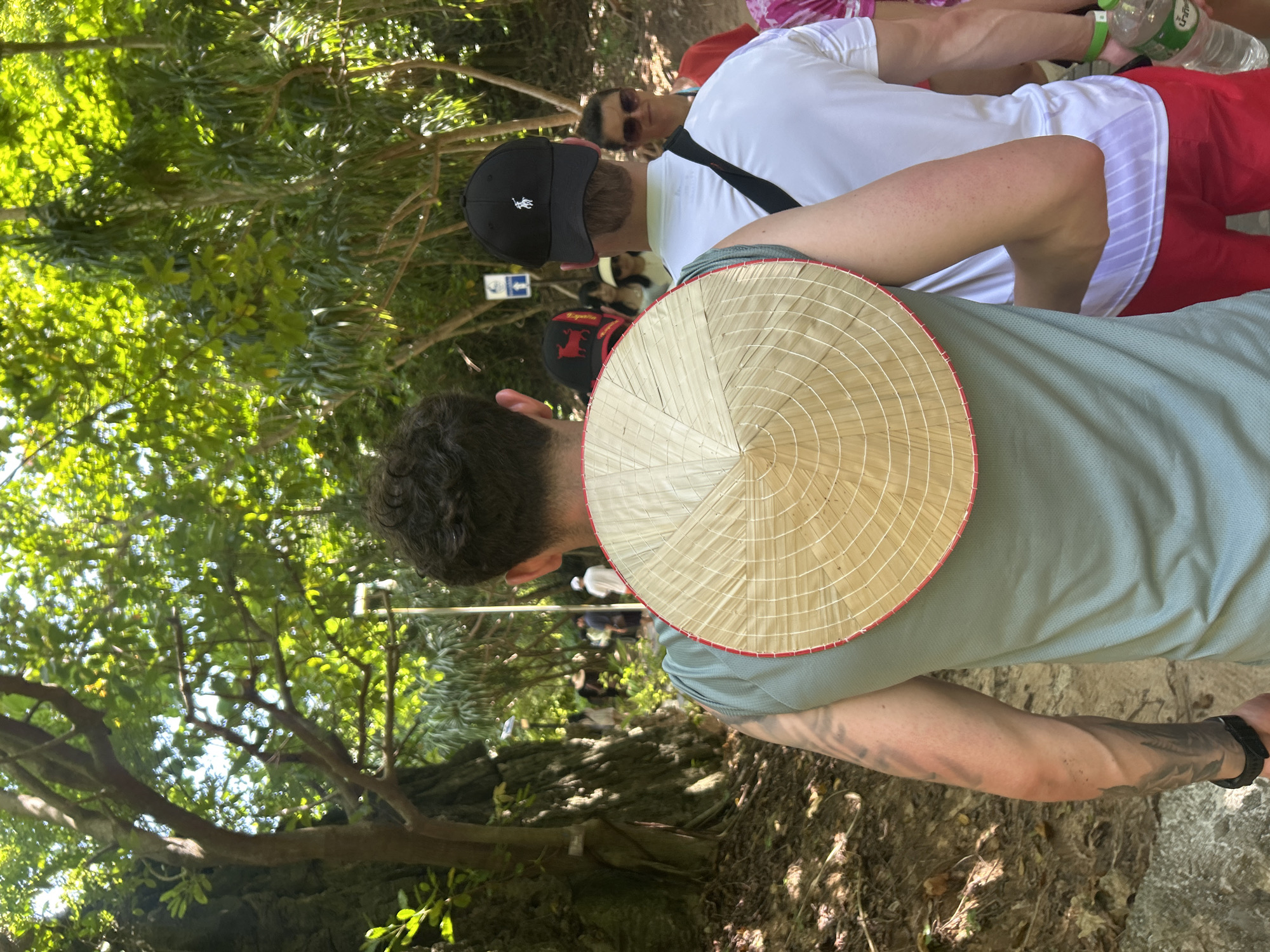 The image captures a group of people walking along a dirt path in a lush, green, outdoor environment, likely a park or nature trail in Ao Nang, Thailand. The foreground is dominated by the back of a person wearing a light blue shirt and a conical Asian hat, which is propped on their back. To their right, another person in a white shirt and red shorts is visible. Further along the path, several other individuals are seen walking, including a person wearing a black cap. The background is filled with dense foliage and trees, suggesting a tropical setting. Sunlight filters through the leaves, creating a dappled effect on the path and surrounding vegetation.