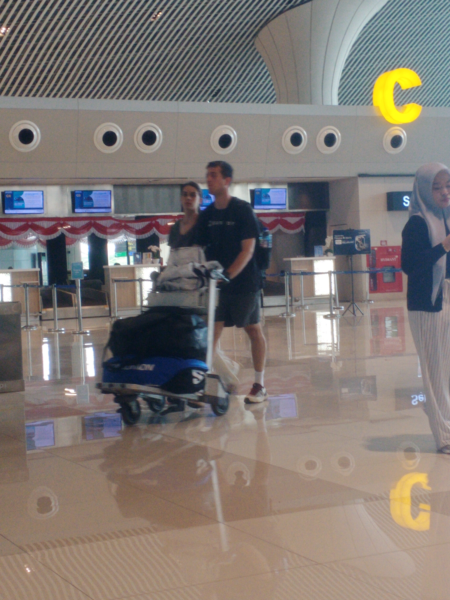 In an indoor airport terminal setting, likely within Baji Mangngai, Indonesia, two individuals are captured in motion. On the left, a man wearing a dark t-shirt and shorts walks alongside a luggage cart piled with a blue duffel bag and other items. He is looking ahead, seemingly focused on his path. To his right, a woman in a dark top and a grey hijab walks behind the luggage cart, her gaze directed downwards. The terminal features a reflective floor, a distinctive white ceiling with angled panels, and wall-mounted screens displaying information. A large, illuminated yellow "C" sign is visible on the upper portion of the wall, serving as a prominent visual element within the scene. The lighting suggests an interior environment with artificial illumination.