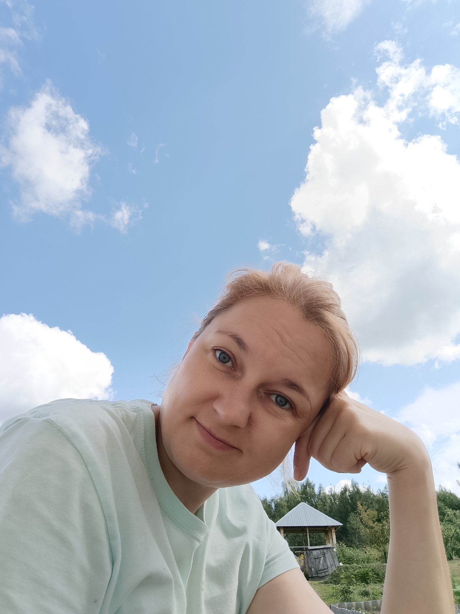 The image is a first-person, slightly downward-angled selfie taken outdoors under a partly cloudy blue sky. The subject is a woman with fair skin and blonde hair, wearing a light blue t-shirt. Her right hand is propped against her face, and she is looking towards the camera with a gentle smile. In the background, there is lush green foliage, a small wooden gazebo with a corrugated metal roof, and more trees. The scene suggests a casual, possibly rural or suburban, outdoor setting. No specific location in Dubovaya, Russia, can be identified from the visual information provided.