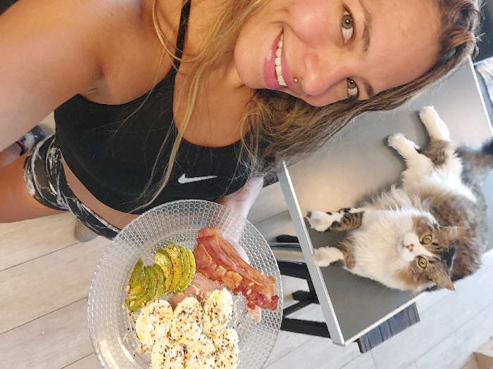 A woman smiles at the camera, holding a plate with avocado slices, bacon, and eggs. She's wearing a black sports bra and patterned shorts. To her right, a fluffy cat with white and brown fur lies on a gray surface, its paws outstretched. The scene is brightly lit, suggesting a daytime setting. The cat stares directly at the camera.